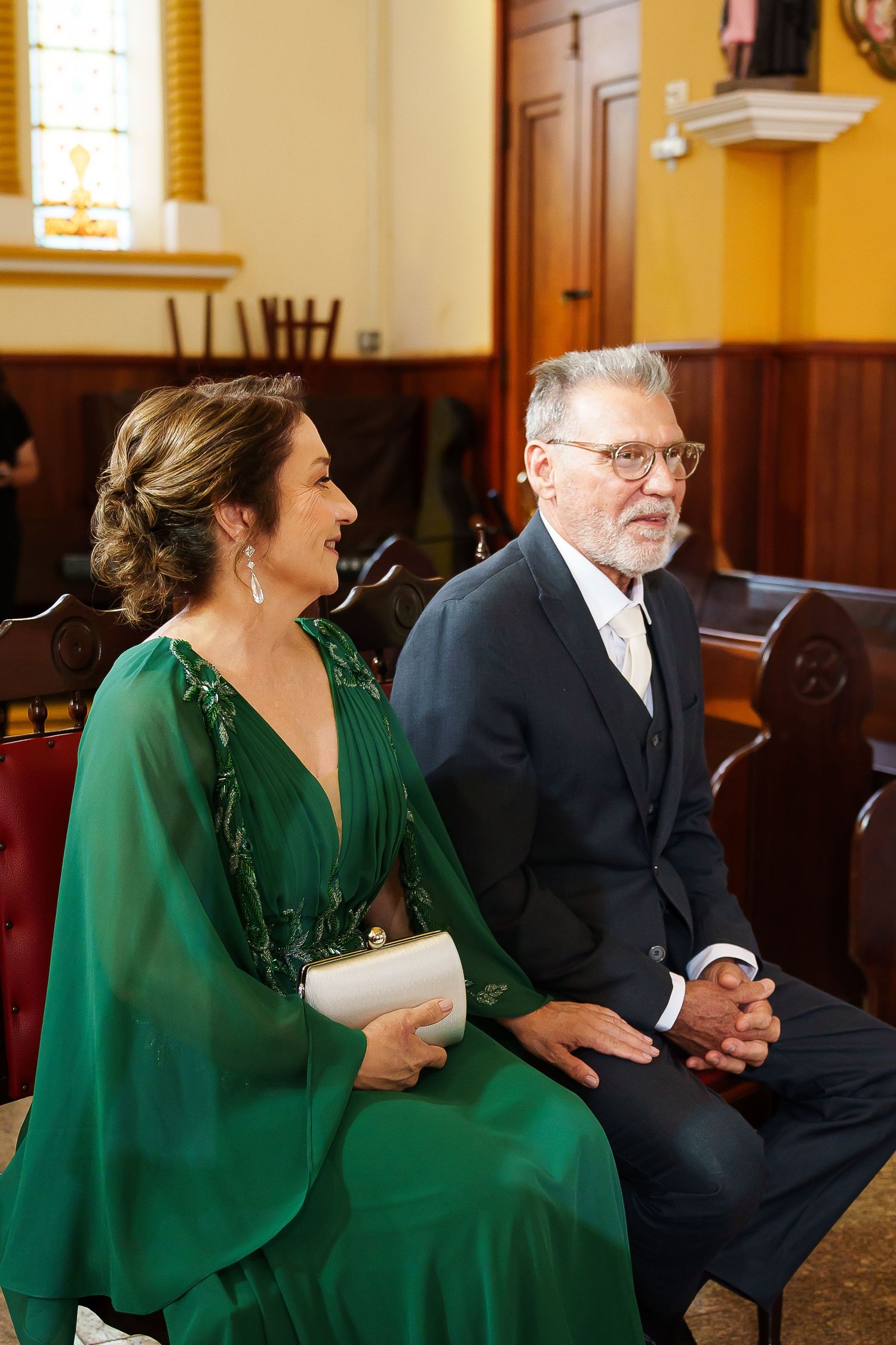 Couple in formal attire seated in church pews, woman in green dress holding clutch, man in navy suit with glasses.