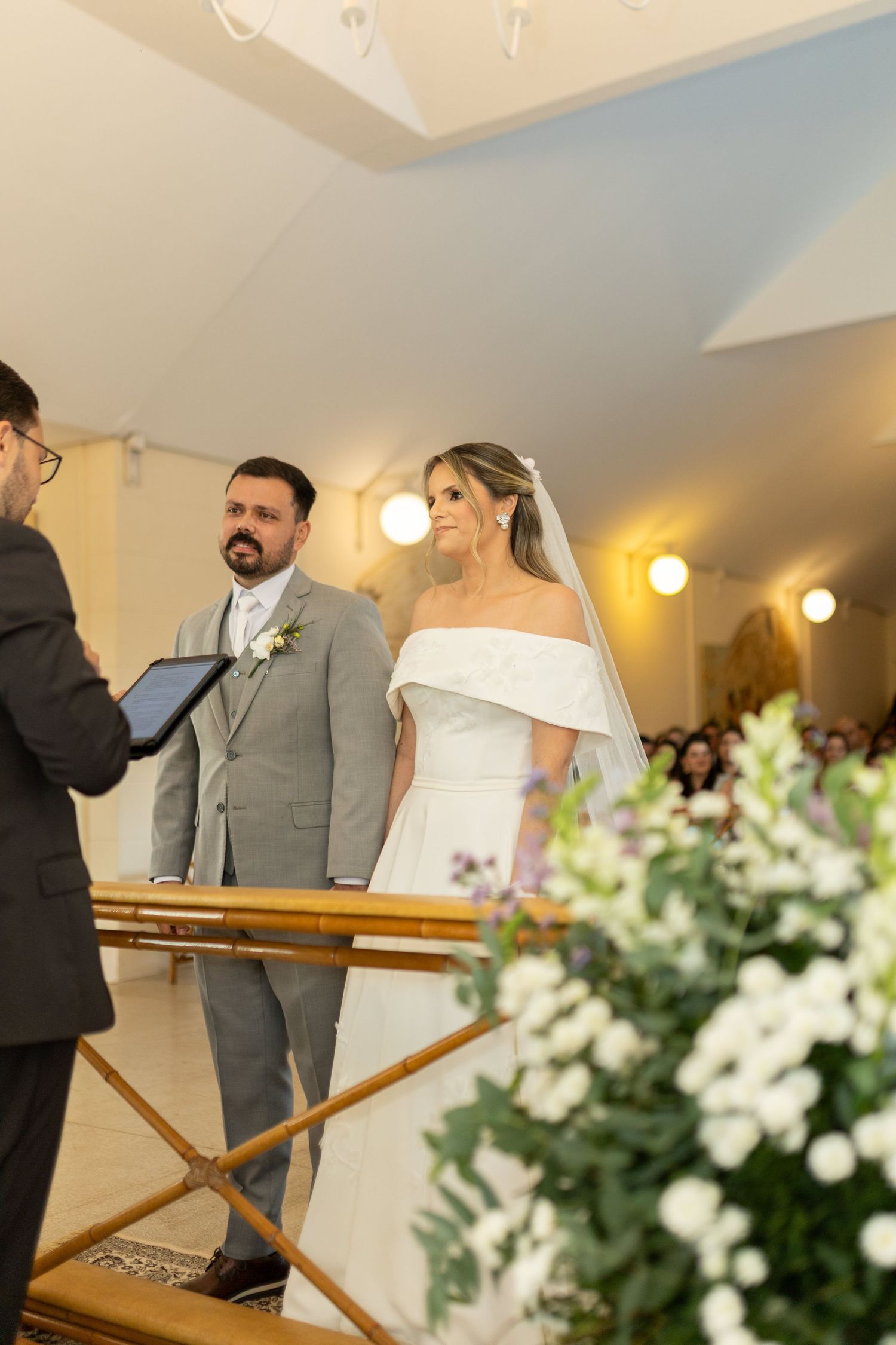 A bride in an off-shoulder white gown and groom in gray suit stand at the altar during their wedding ceremony.