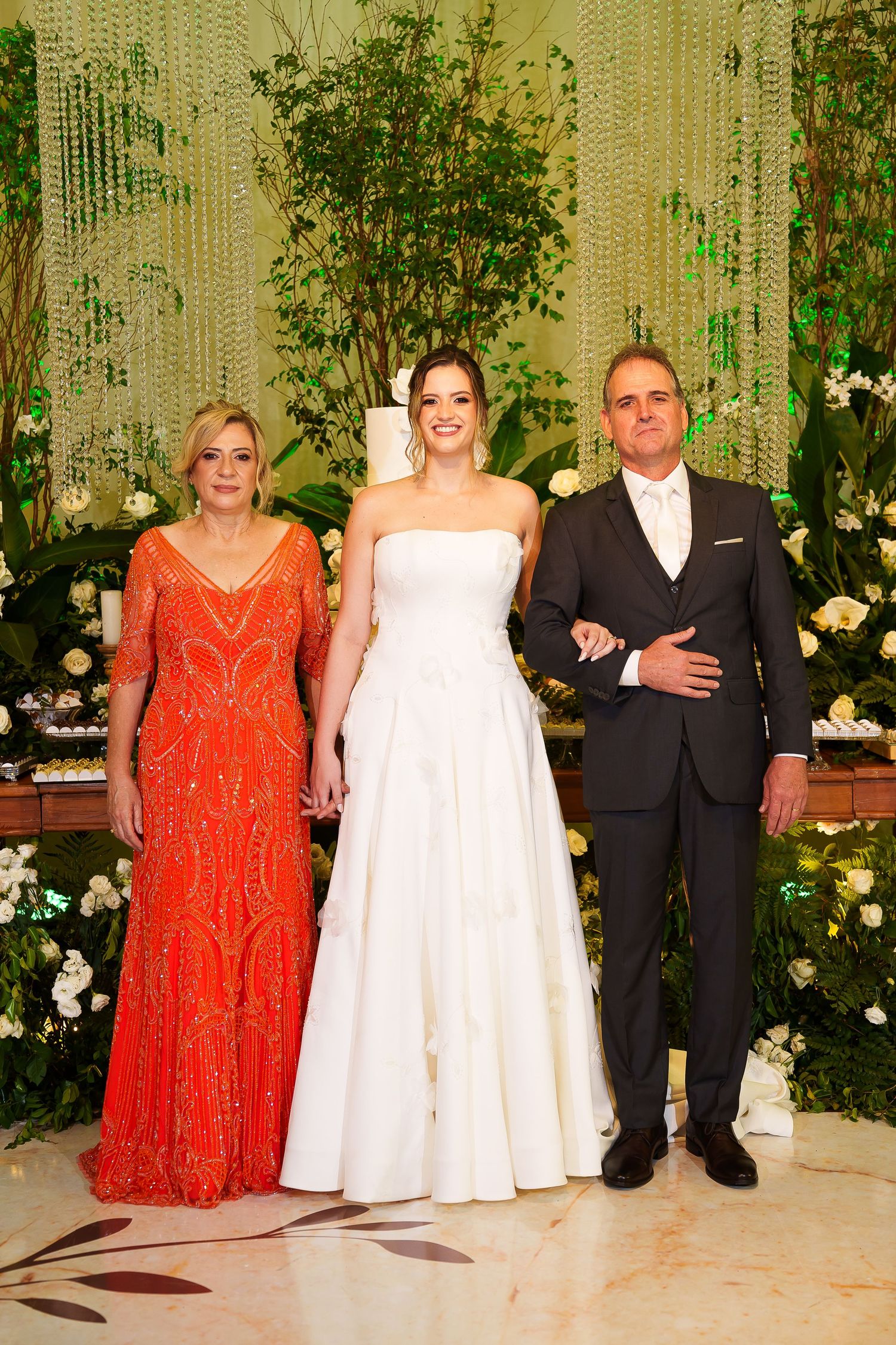 Bride in white gown poses with parents, mother in orange beaded dress, father in dark suit, at elegant floral wedding venue.