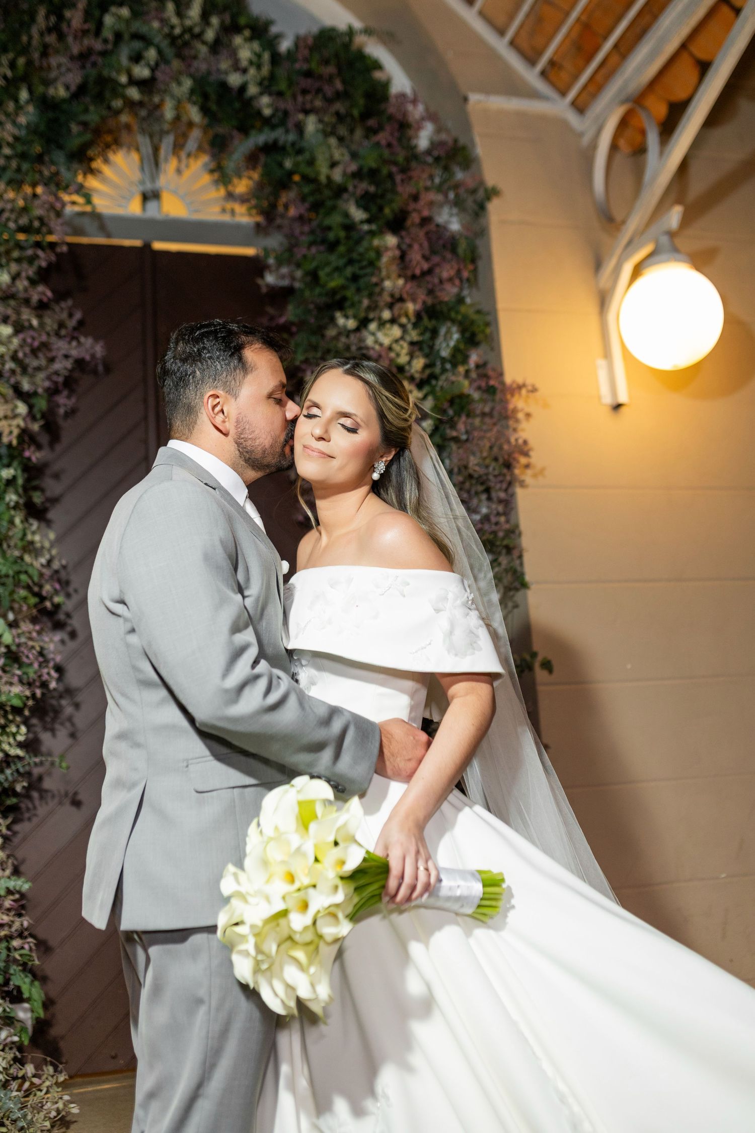 Bride in white off-shoulder gown holding calla lily bouquet, embraced by groom in gray suit at elegant floral venue.