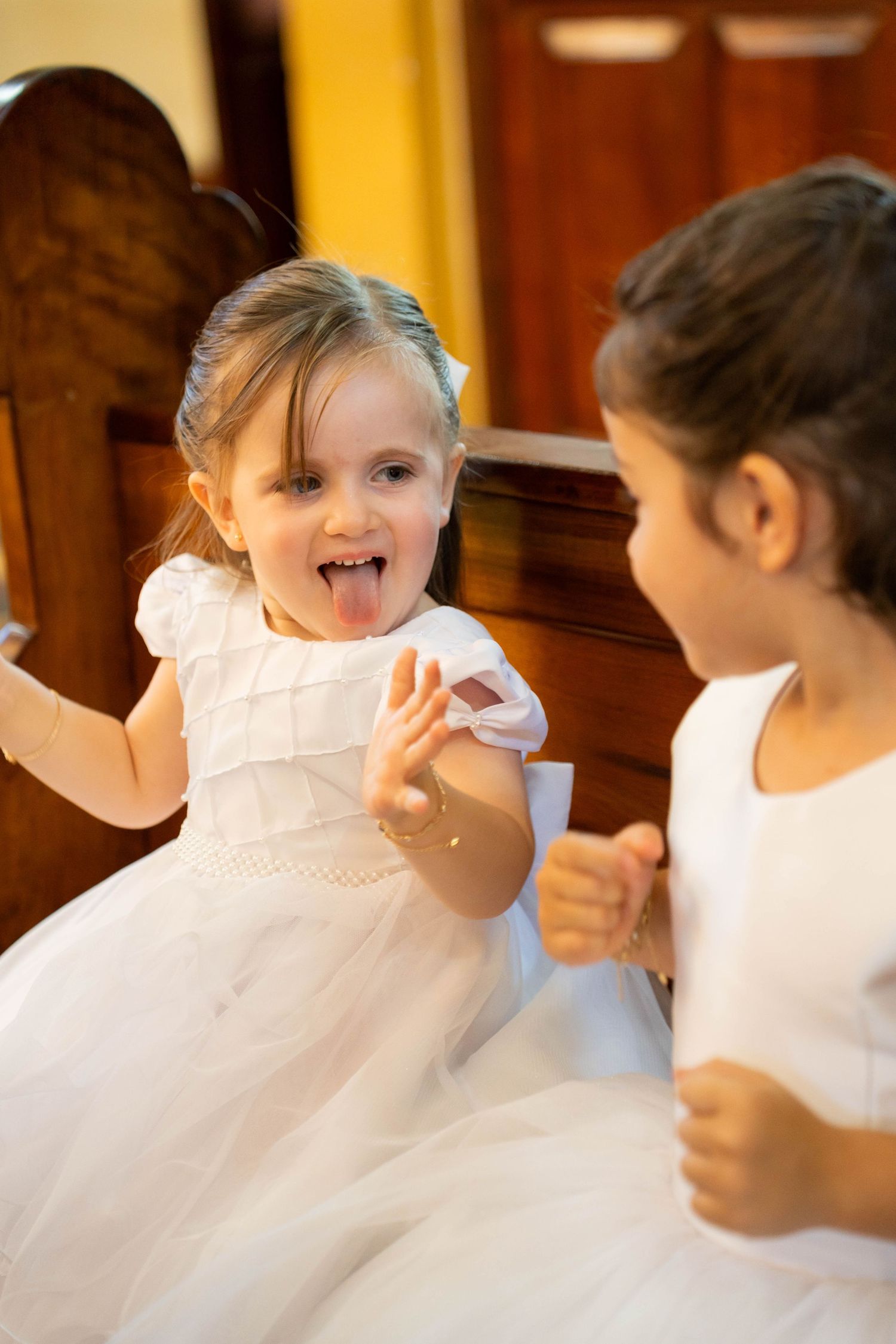 Two young girls in white dresses playfully interact, one sticking out her tongue and reaching toward the other.