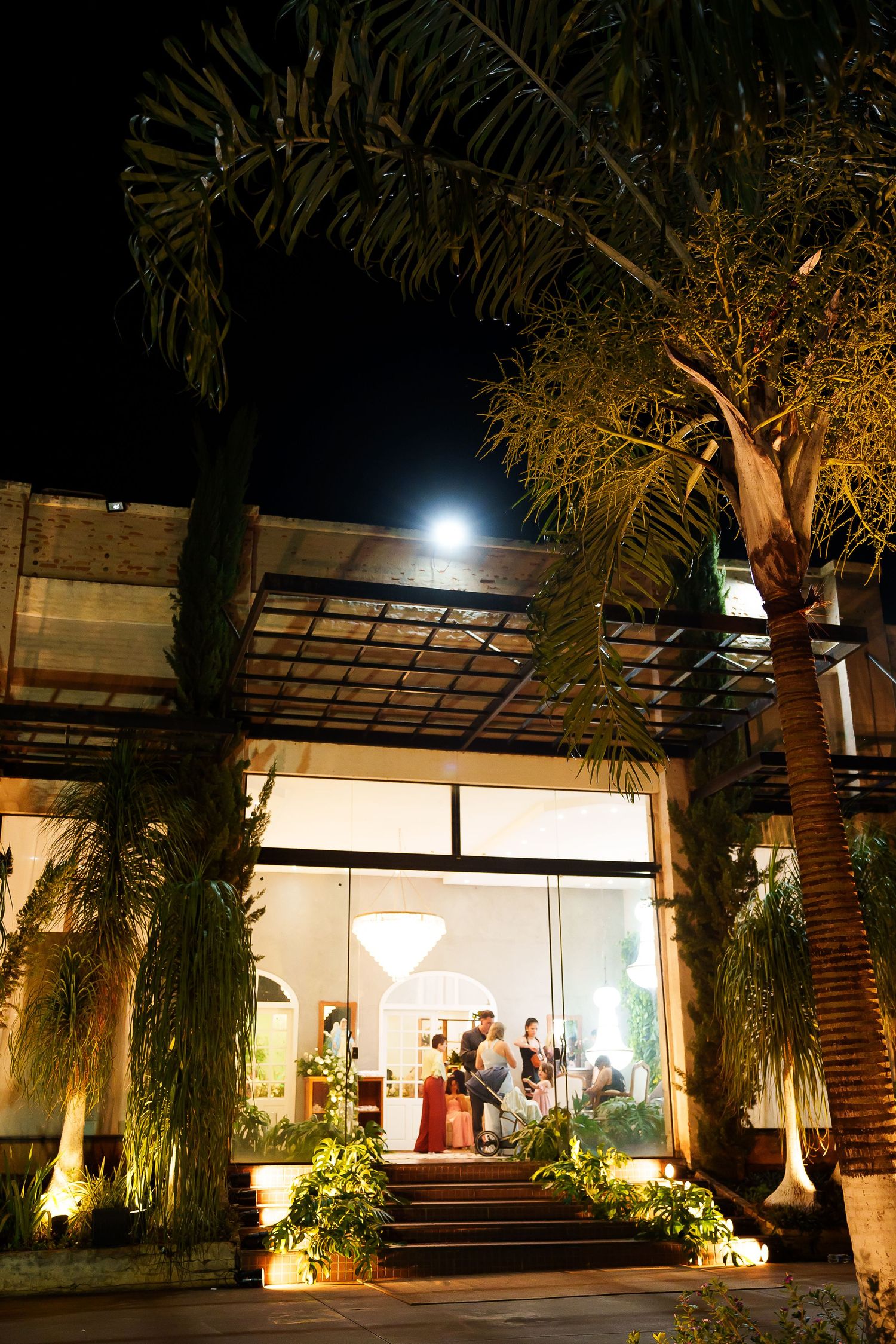Elegant outdoor venue entrance at night with tropical trees, steps, and guests visible through large glass windows.