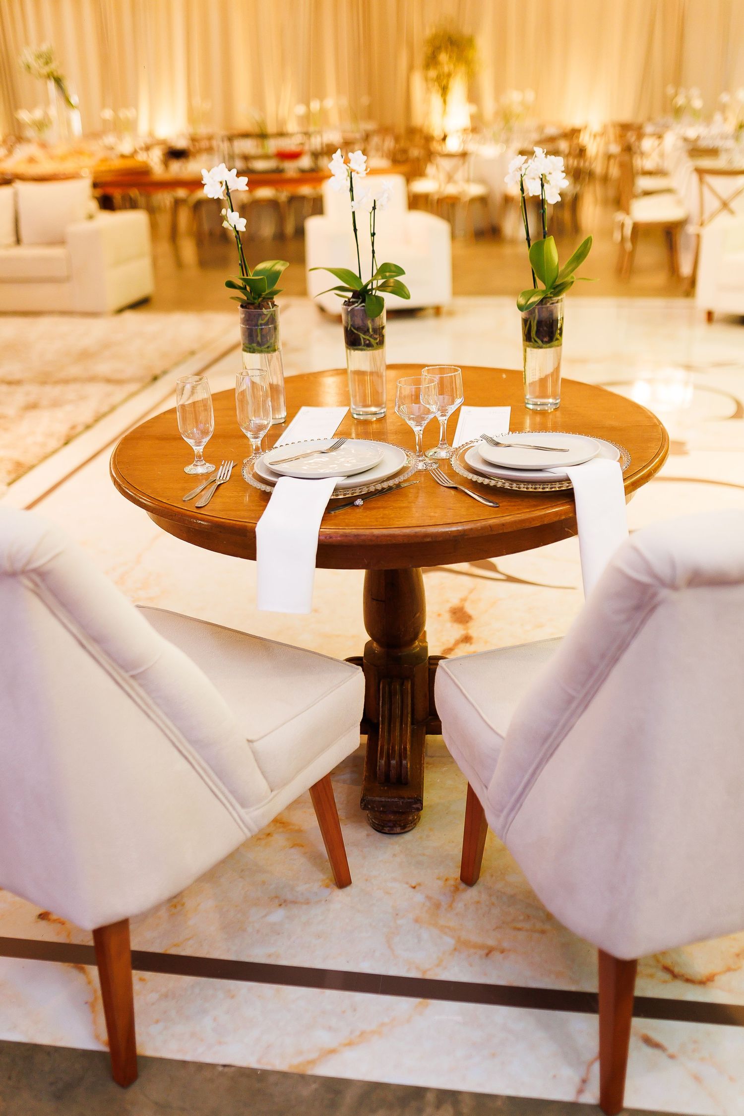 Elegant wedding reception table set for two with white orchids, crystal glasses, and ivory chairs in a luxurious ballroom.