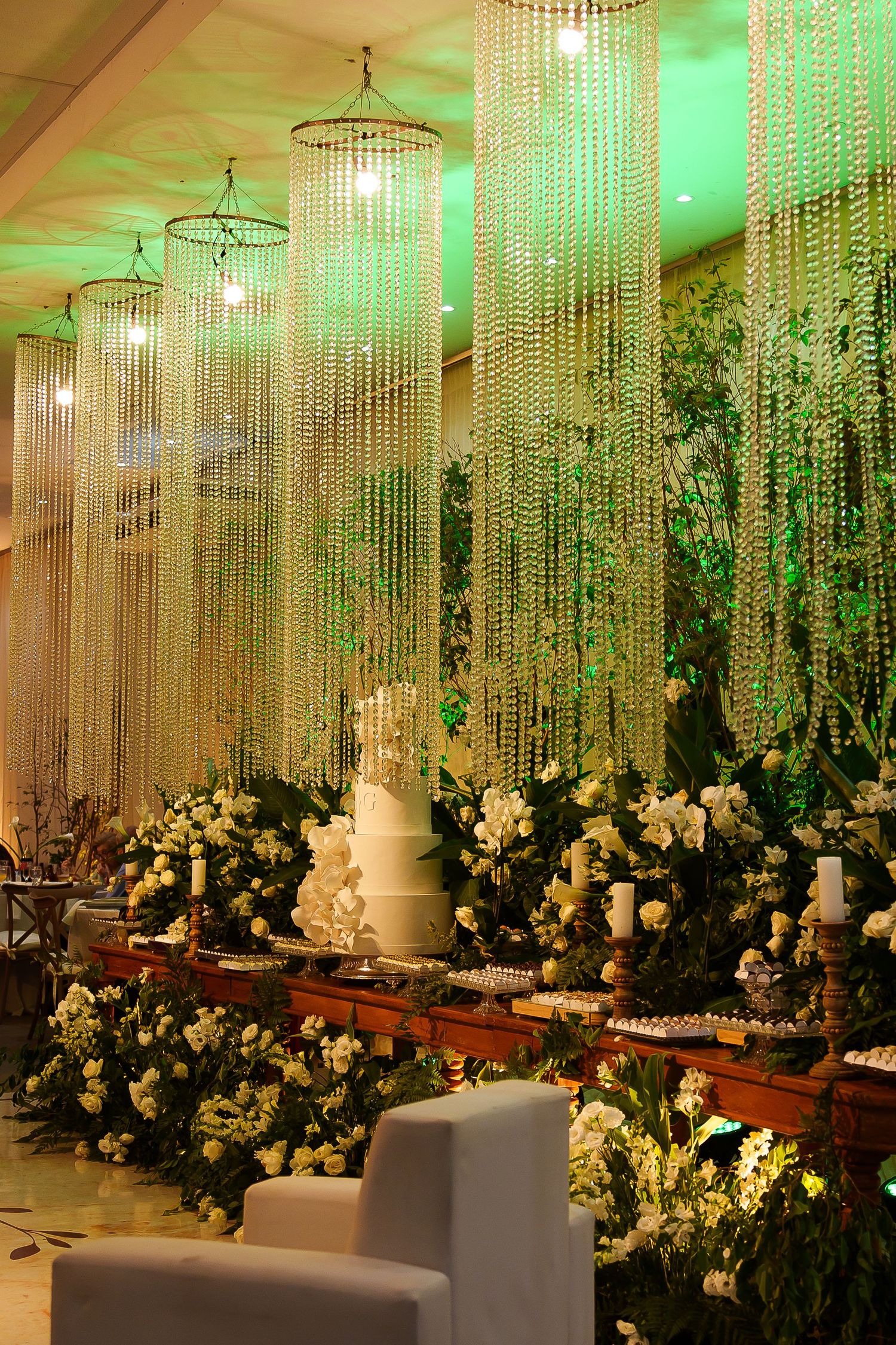 Elegant wedding reception with crystal chandelier curtains, white tiered cake, and lush floral arrangements in green-lit venue.