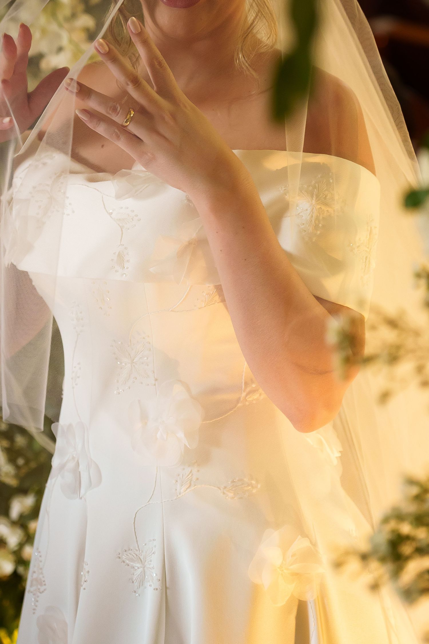Bride in white embroidered wedding gown with veil, hands gracefully posed, surrounded by soft golden light and flowers.