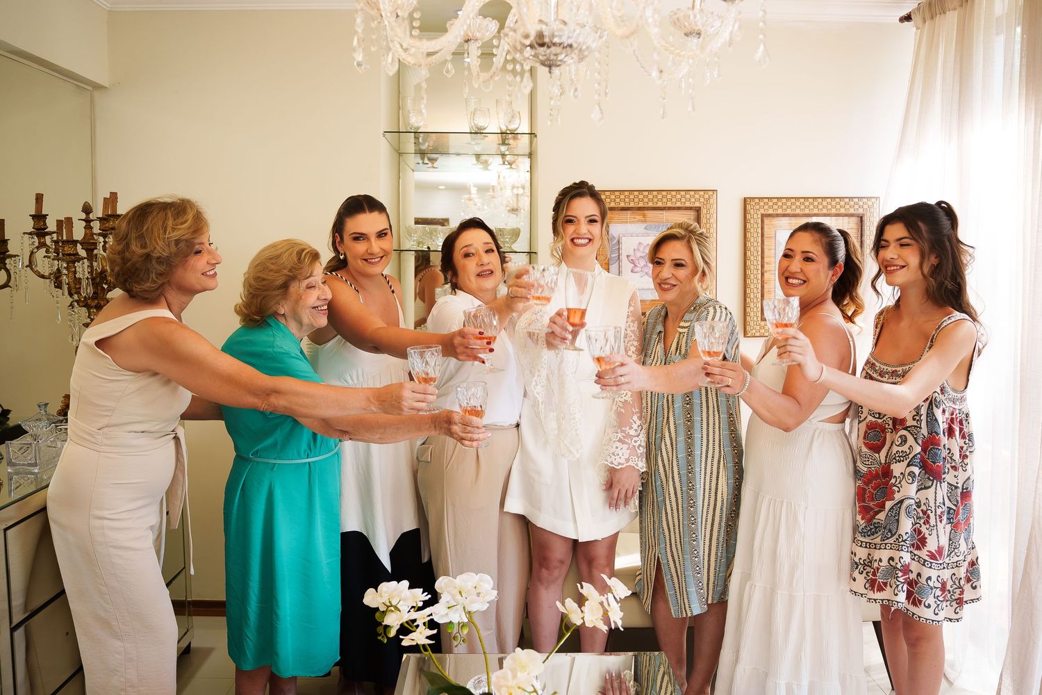 Bride and bridesmaids toasting with champagne glasses in elegant white room during wedding preparations.