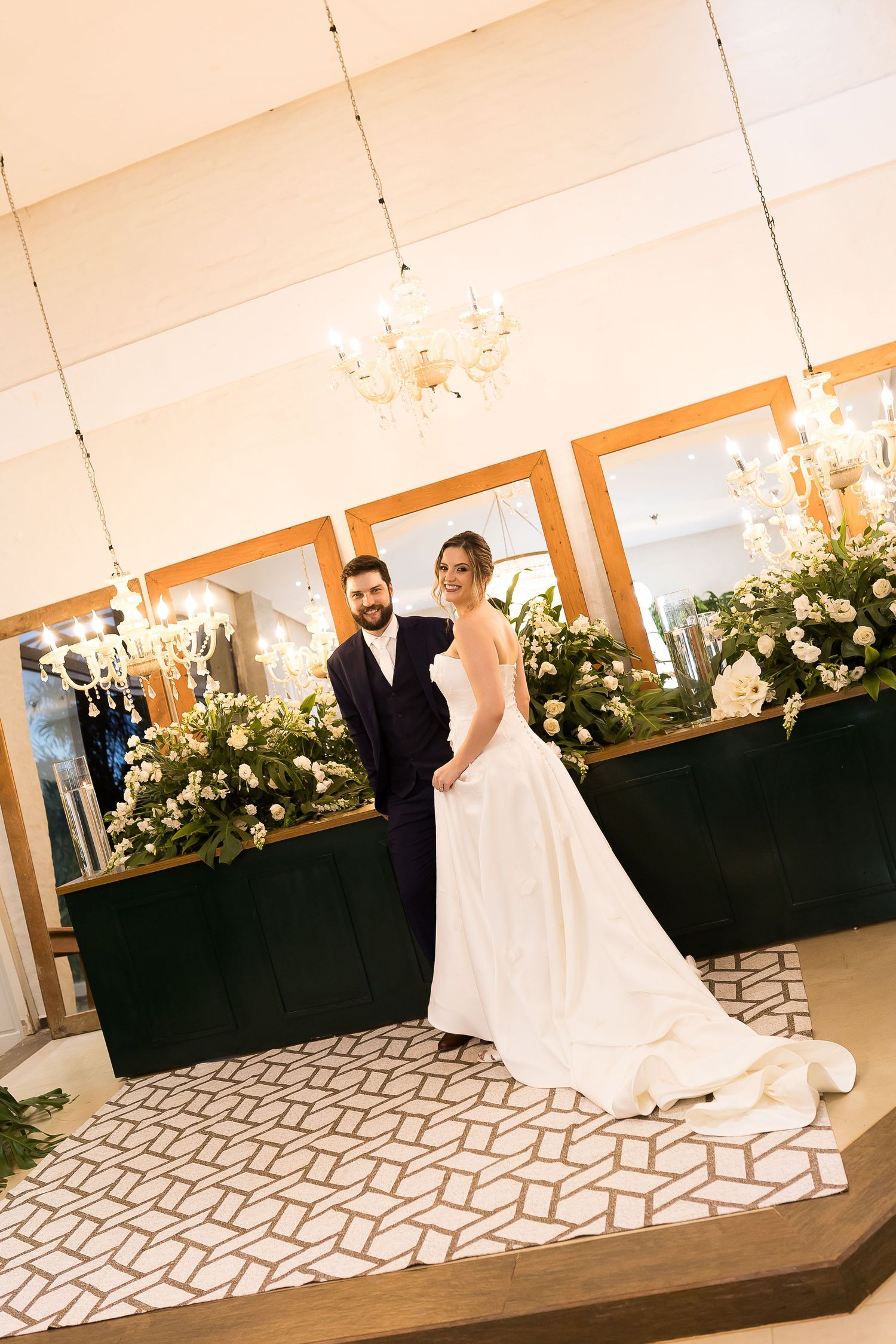 Bride in white gown and groom in navy suit pose elegantly in a floral-decorated wedding venue with chandeliers.