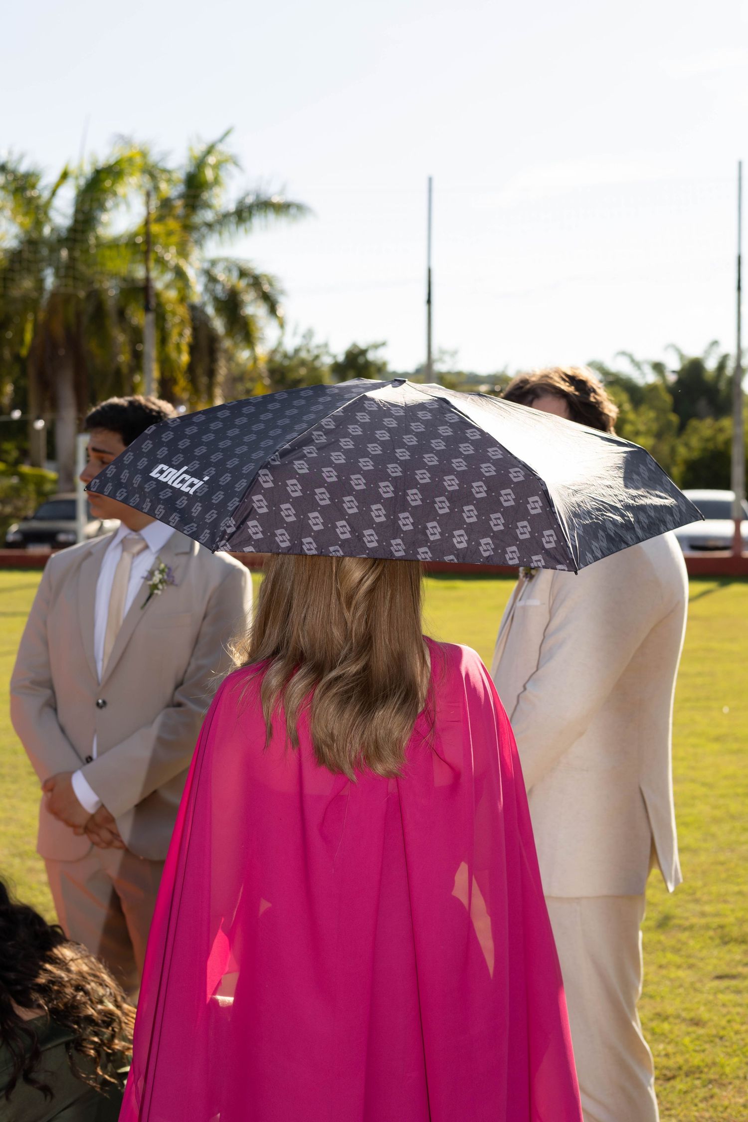 Woman in pink cape holding patterned umbrella at outdoor wedding ceremony with palm trees.
