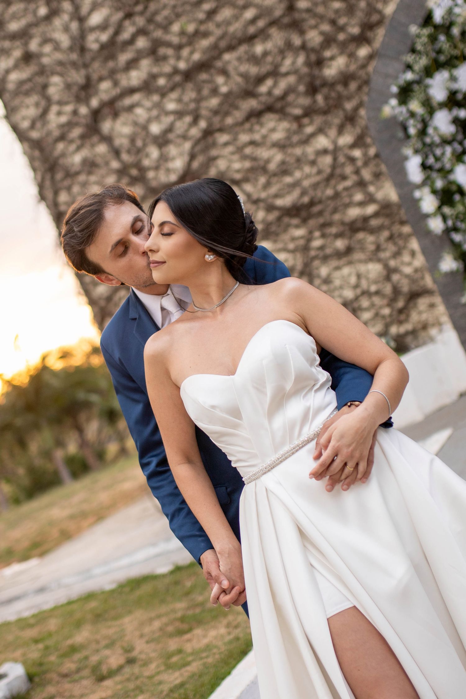 Bride in white strapless gown and groom in blue suit share romantic moment outdoors at sunset.