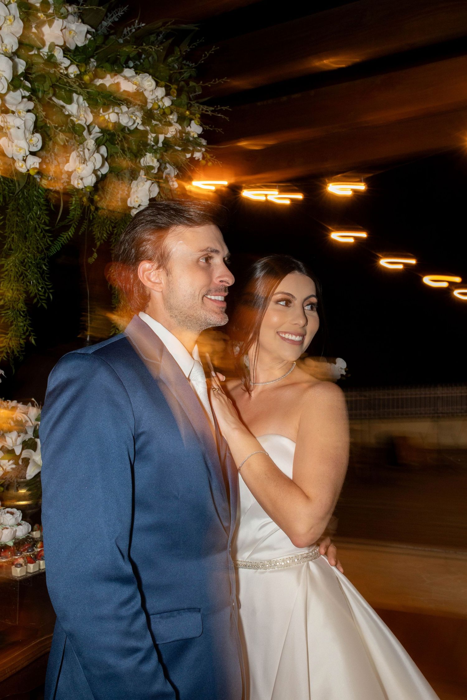 Bride and groom smiling together at elegant evening wedding reception with floral arch and warm string lights.