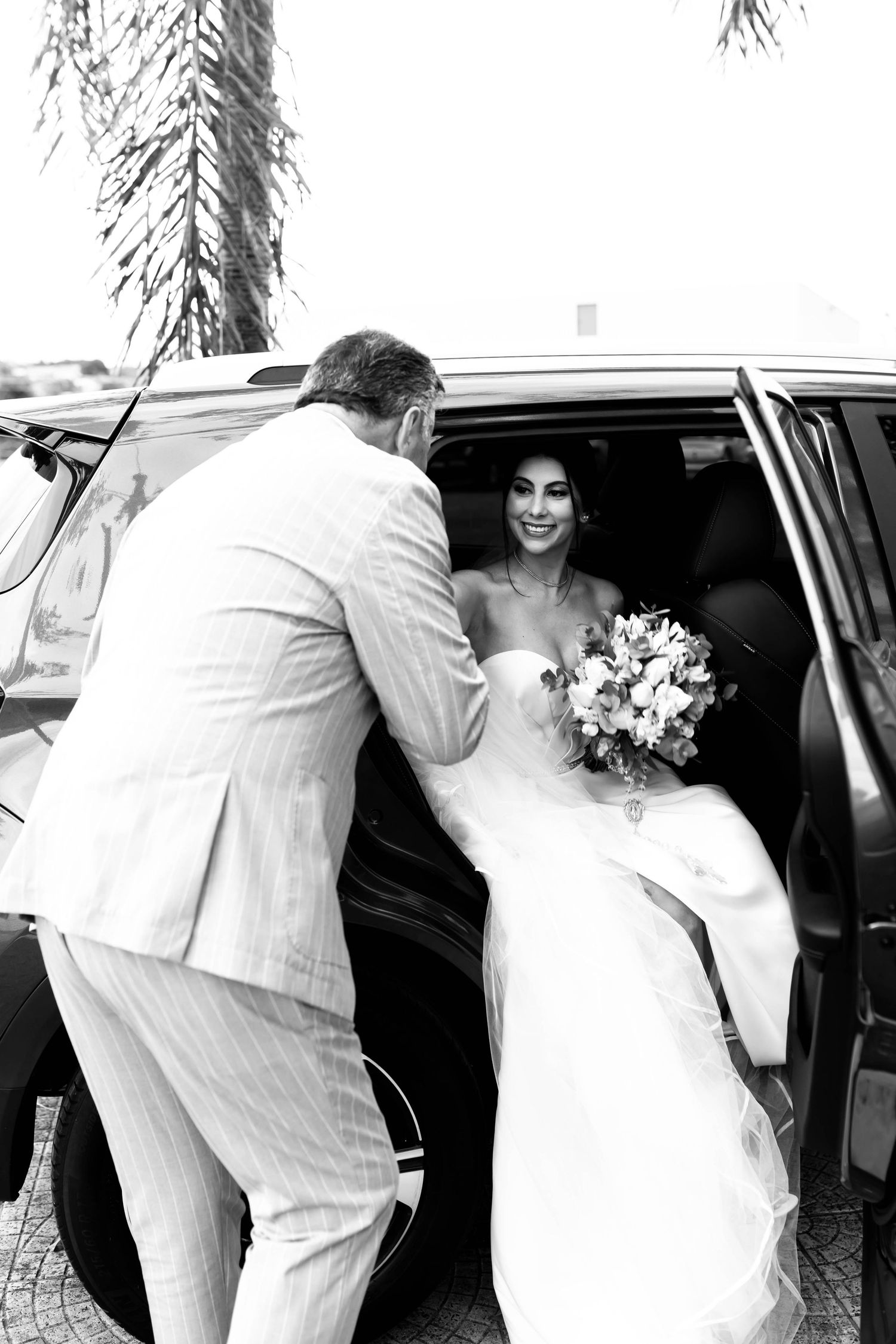 Groom in white suit helping smiling bride exit car while she holds her wedding bouquet.
