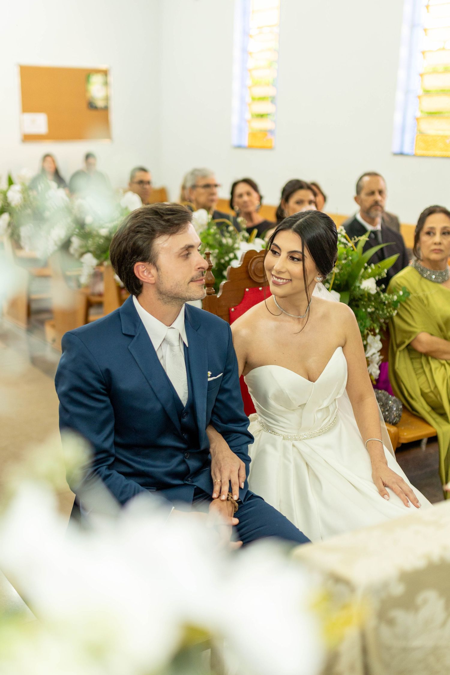 A bride and groom sit together at their church wedding ceremony, smiling lovingly at each other, surrounded by floral arrangements.