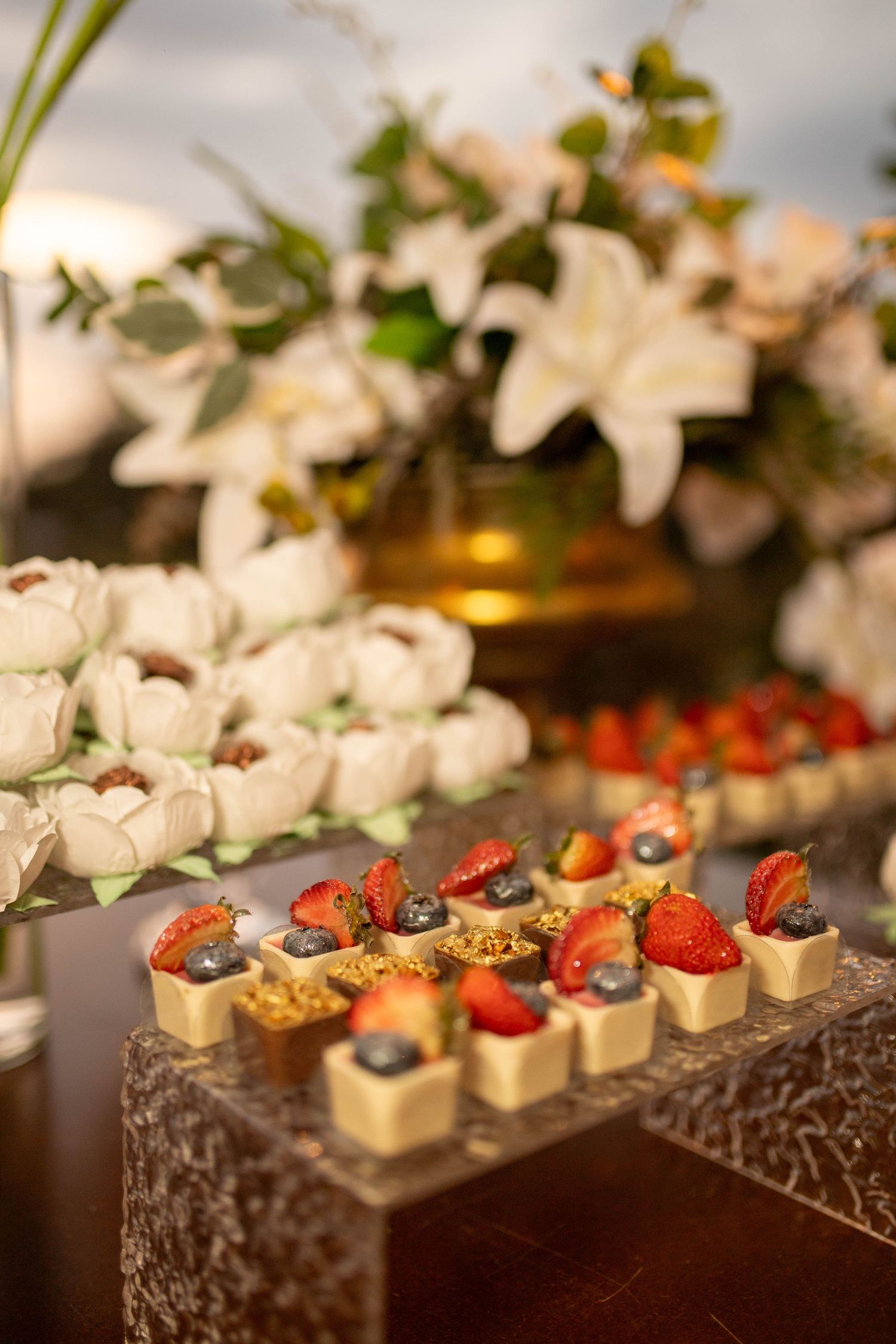 Elegant dessert display with fruit-topped chocolate cups, macarons, and white floral arrangement at a wedding reception.