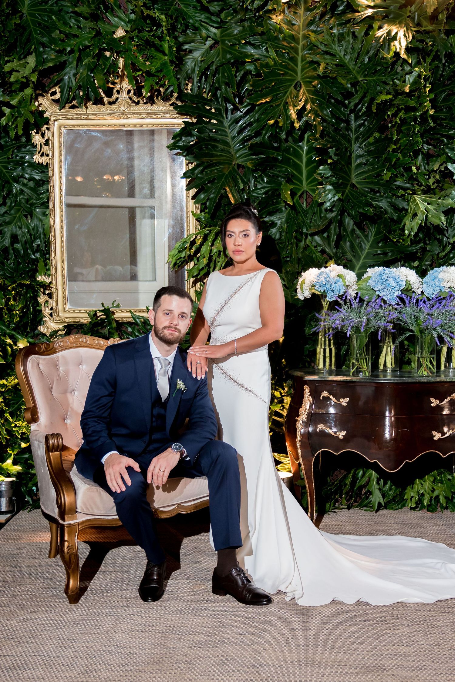Bride in white gown stands beside groom in navy suit seated in gold chair, surrounded by lush greenery.