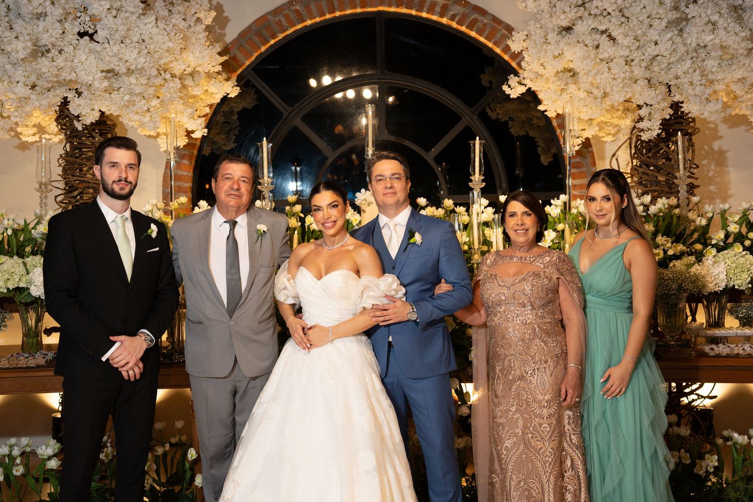 Bride and groom pose with family members in elegant wedding venue decorated with white floral arrangements.