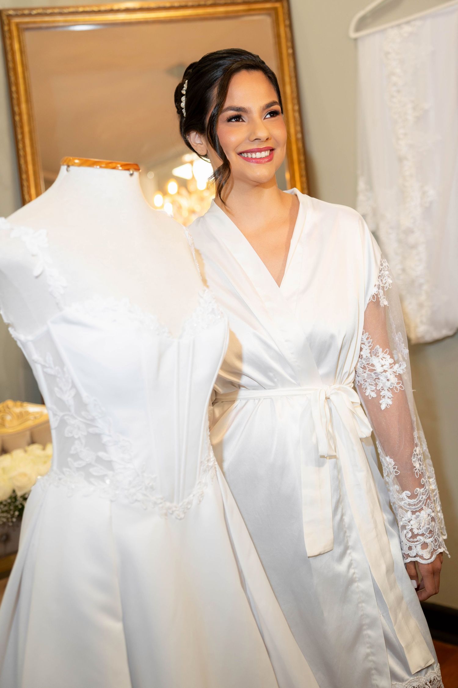 Smiling bride in white satin robe with lace sleeves stands beside a mannequin displaying an elegant wedding gown in a bridal boutique.