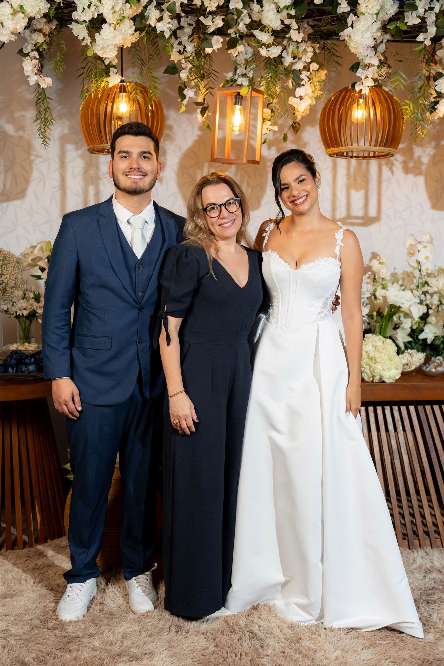 A couple in wedding attire poses with a guest under floral decorations and warm pendant lights at an elegant wedding venue.