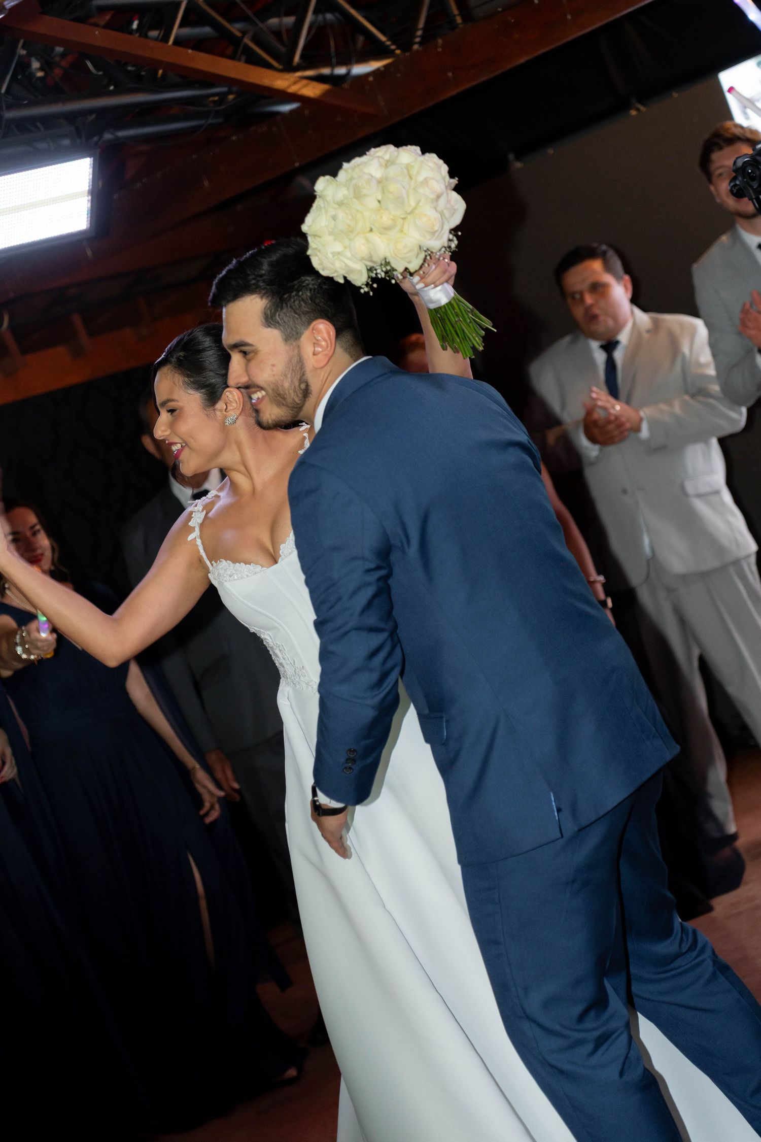 Bride and groom share a romantic dance at their wedding reception, bride holding white rose bouquet, guests celebrating in background.
