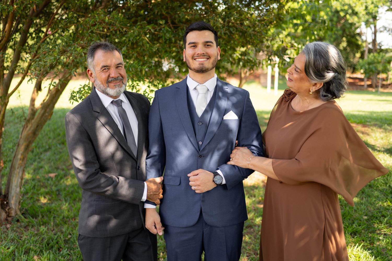 A groom in a navy suit stands smiling between his parents outdoors, surrounded by lush green trees.