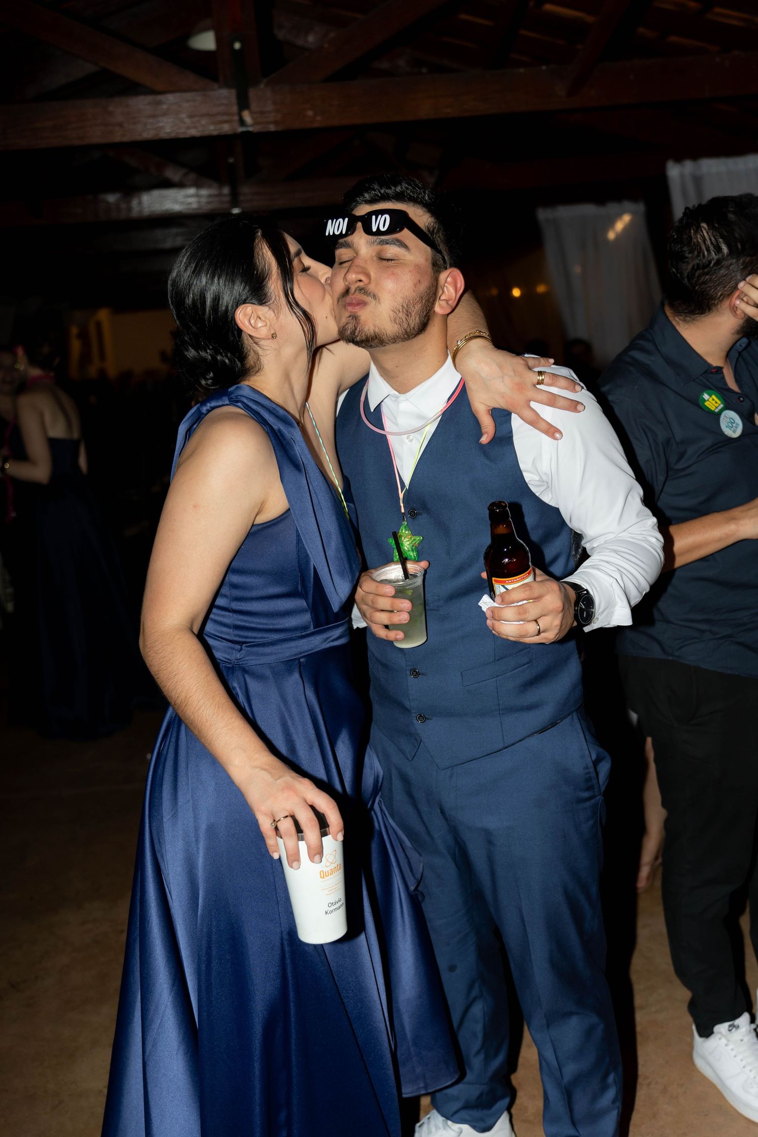 Couple in navy blue attire sharing a kiss at a wedding reception, holding drinks on the dance floor.