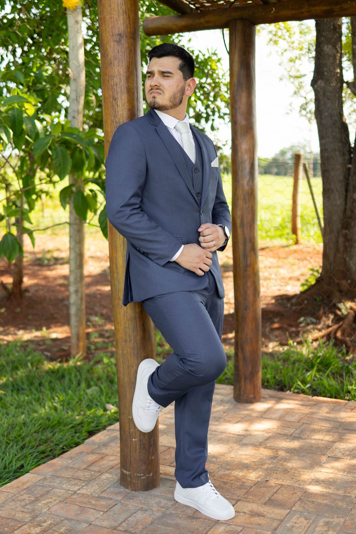Man in navy blue three-piece suit and white sneakers leaning against wooden post in garden setting.