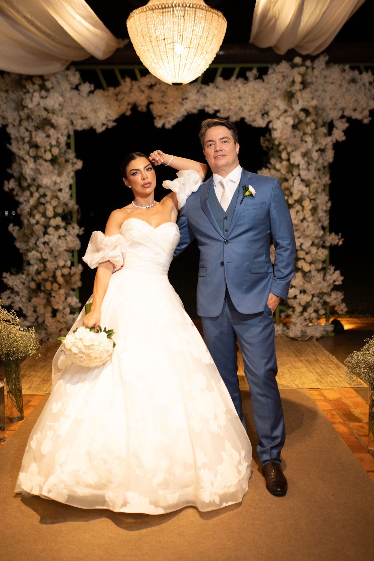 Bride in white ball gown and groom in blue suit pose together at elegant floral wedding ceremony.