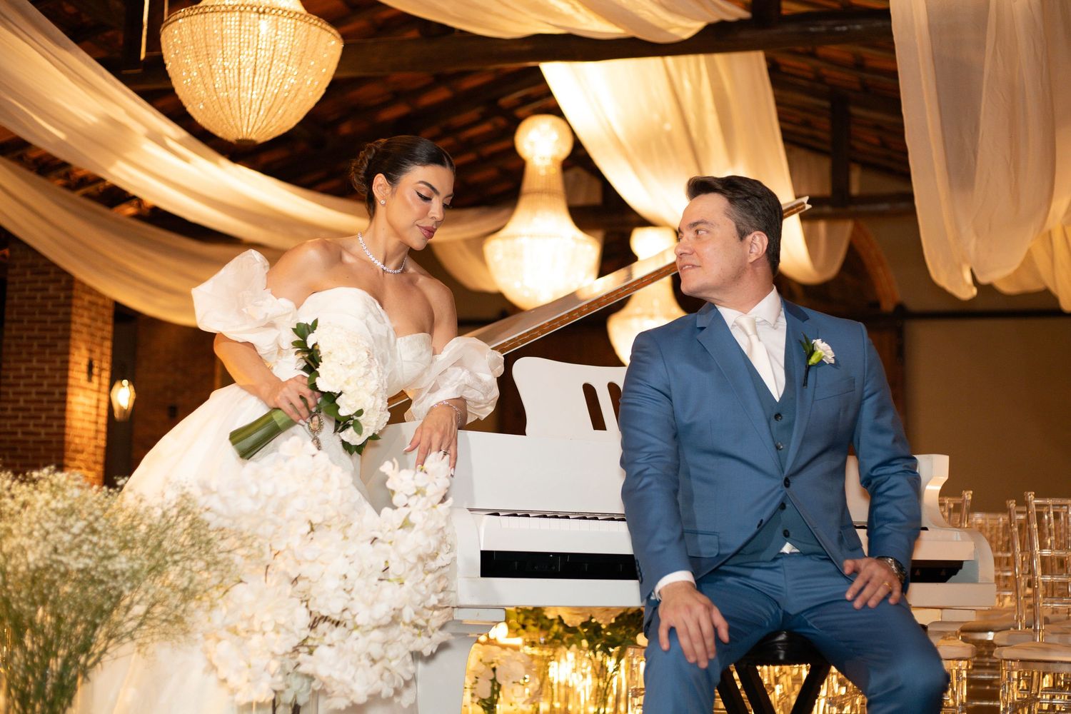 Elegant bride in white gown and groom in blue suit share a romantic moment by a white piano at their wedding reception.