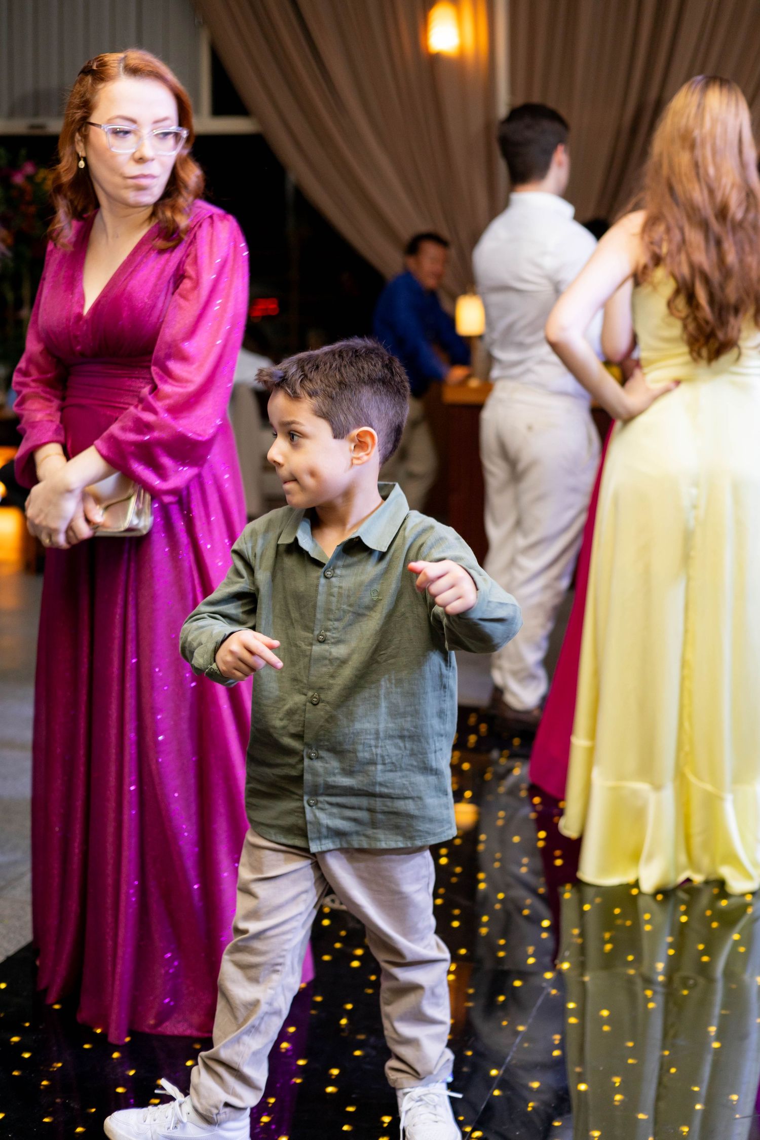 A young boy dances at a formal event beside a woman in a pink gown, with guests in elegant attire in the background.