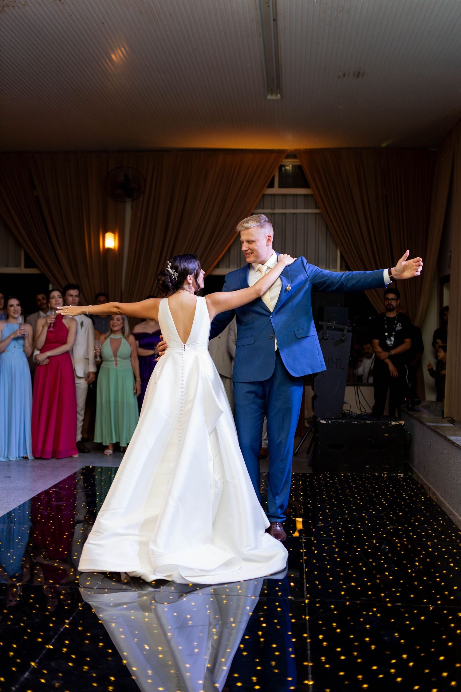 Bride and groom share their first dance on a starlit floor at their wedding reception, guests watching in background.