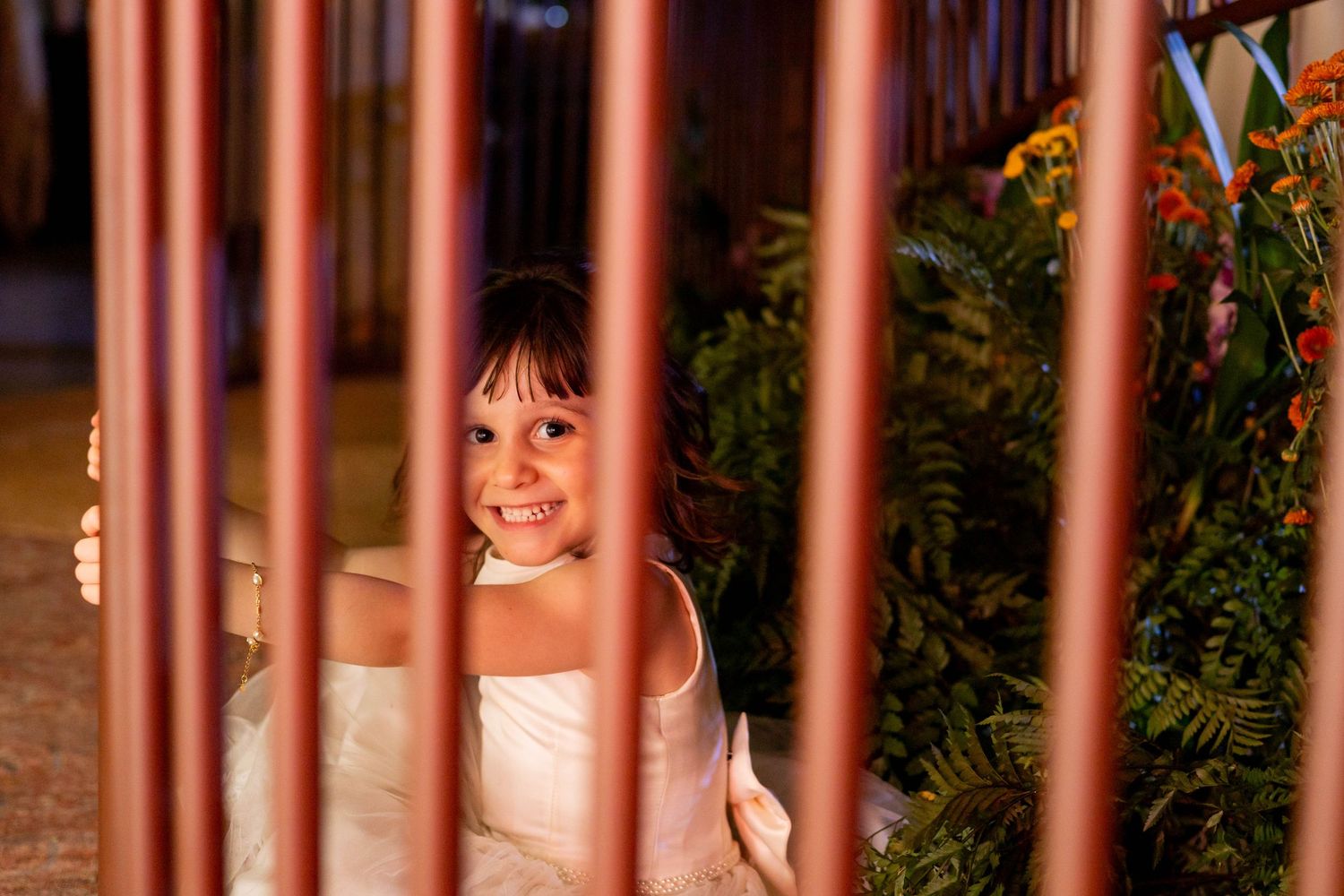 A smiling young girl in a white dress peers through decorative copper bars surrounded by lush greenery.