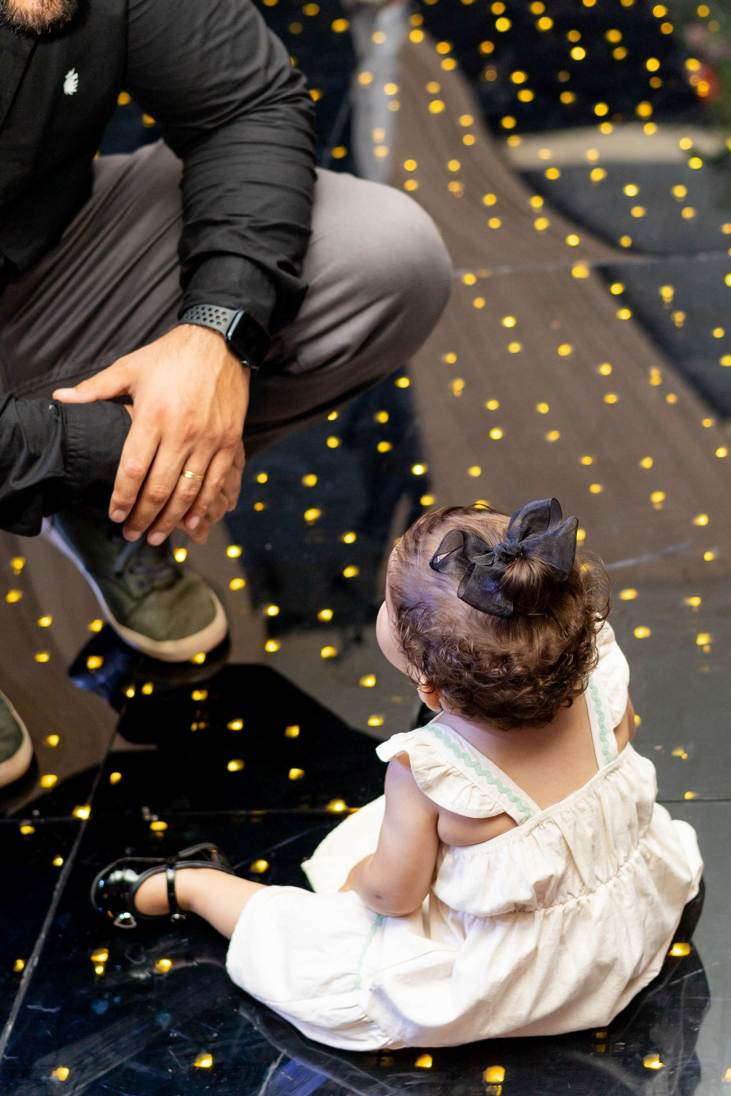 A baby girl in white dress sits on dark floor with golden bokeh lights beside a crouching man.