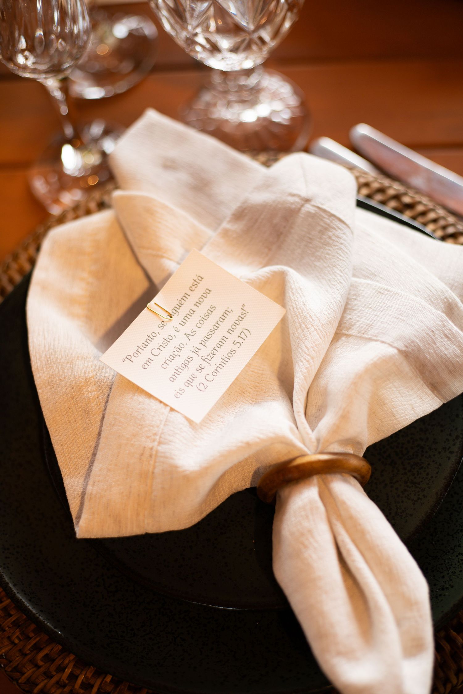 Elegant table setting with white linen napkin, gold ring, and a small card on a dark plate with crystal glasses.
