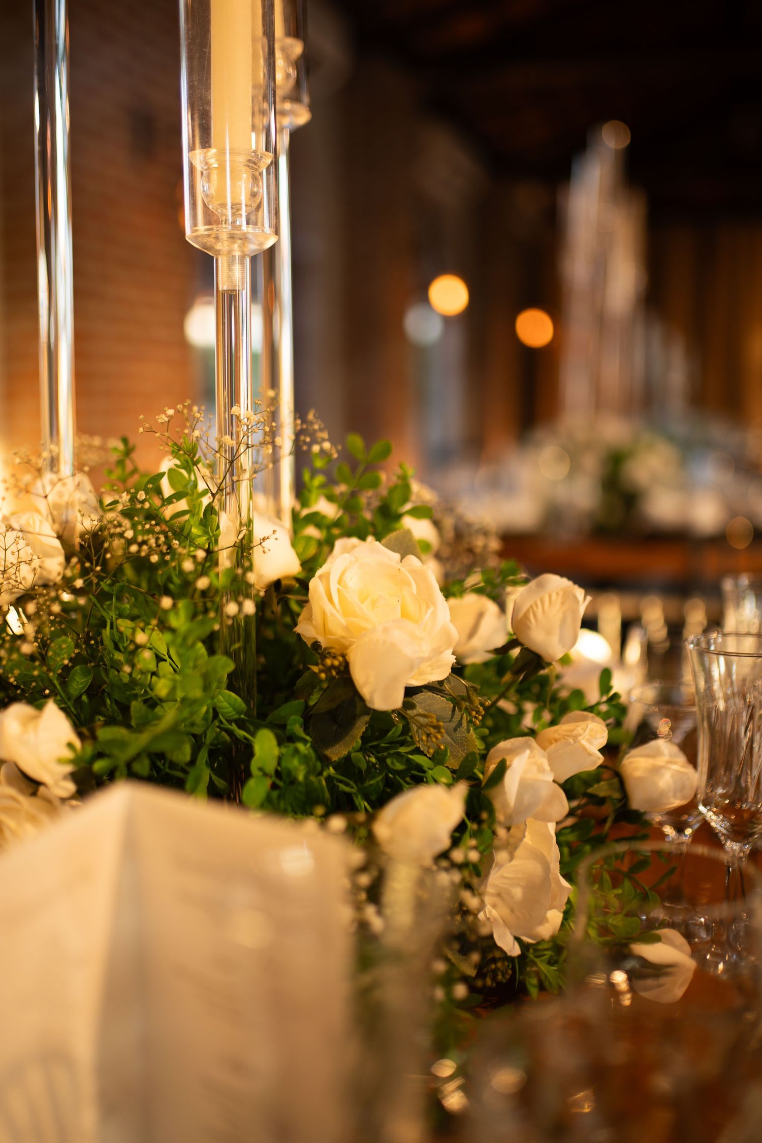 Elegant wedding table with white roses, greenery centerpiece, and tall glass candle holders in a warmly lit venue.