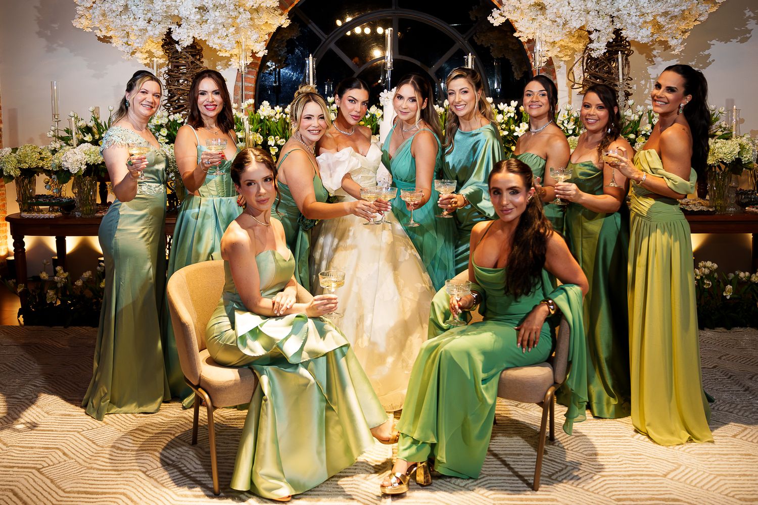 Bride in white gown poses with bridesmaids in green satin dresses at elegant floral wedding reception.