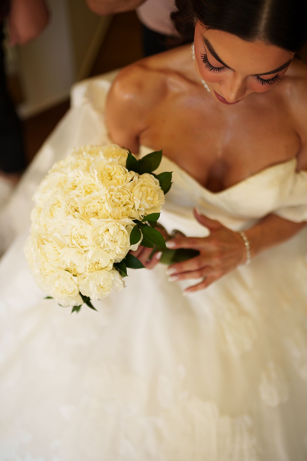 Bride in strapless white ball gown holding elegant bouquet of cream roses with green leaves, looking down at flowers.