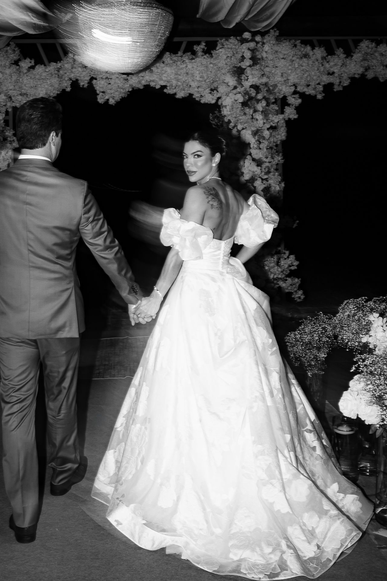 Black and white photo of a bride in a backless ball gown holding hands with groom at elegant wedding reception.