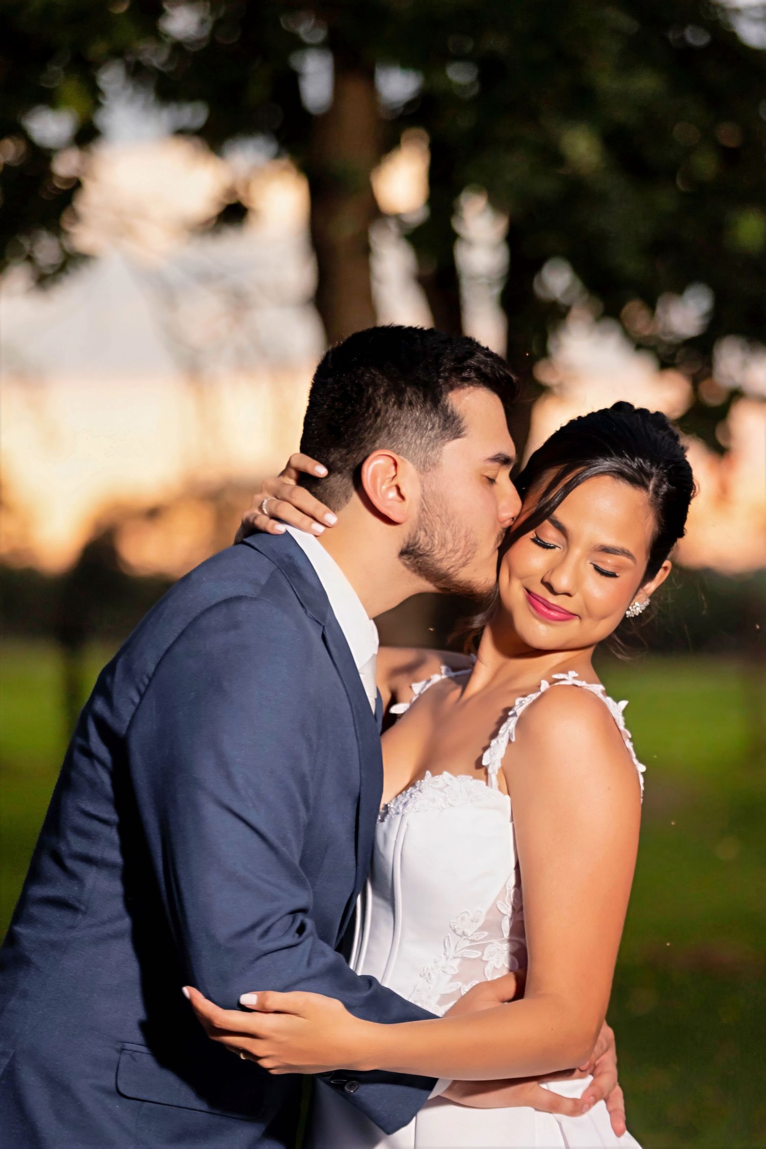 Groom in blue suit kissing bride on cheek outdoors at golden hour, bride smiling in white wedding dress.