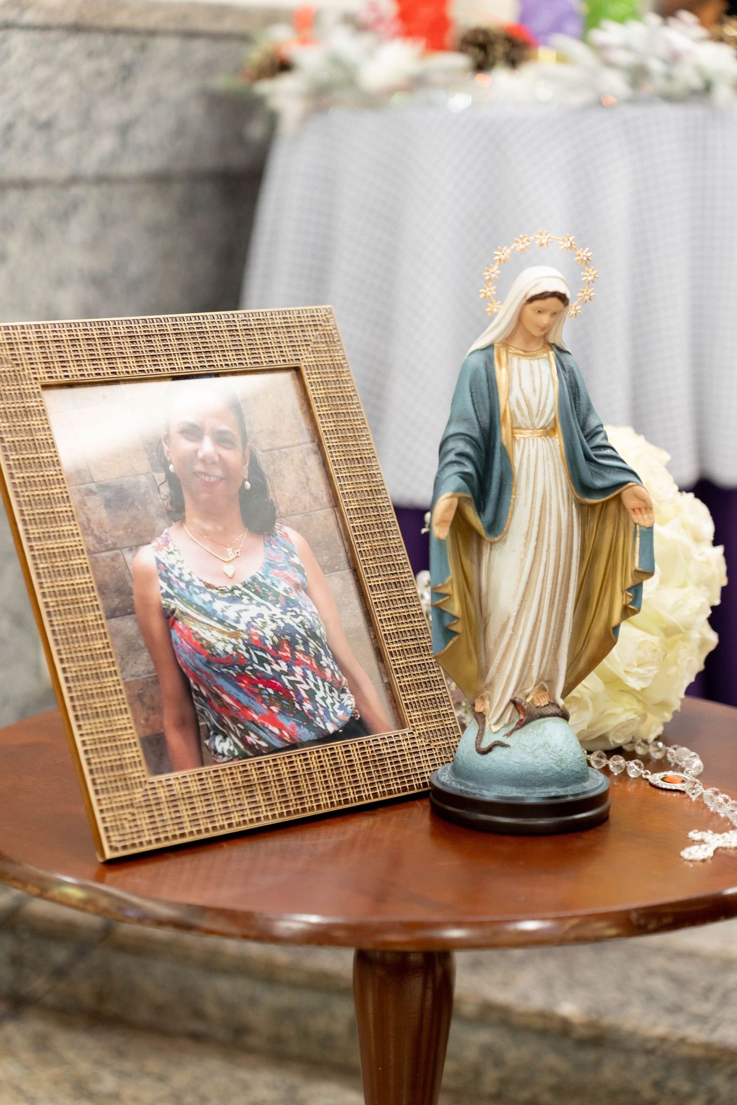 A framed photo of a woman displayed beside a Virgin Mary statue with rosary beads on a wooden table.