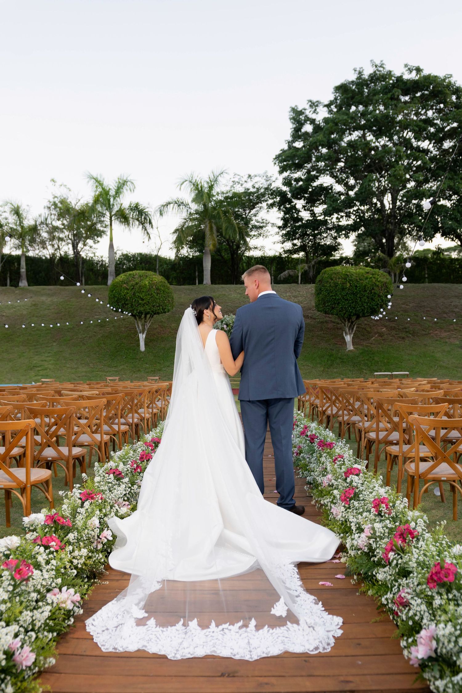 Bride and groom stand together at outdoor wedding aisle lined with floral arrangements and wooden chairs, bride wearing long lace veil.