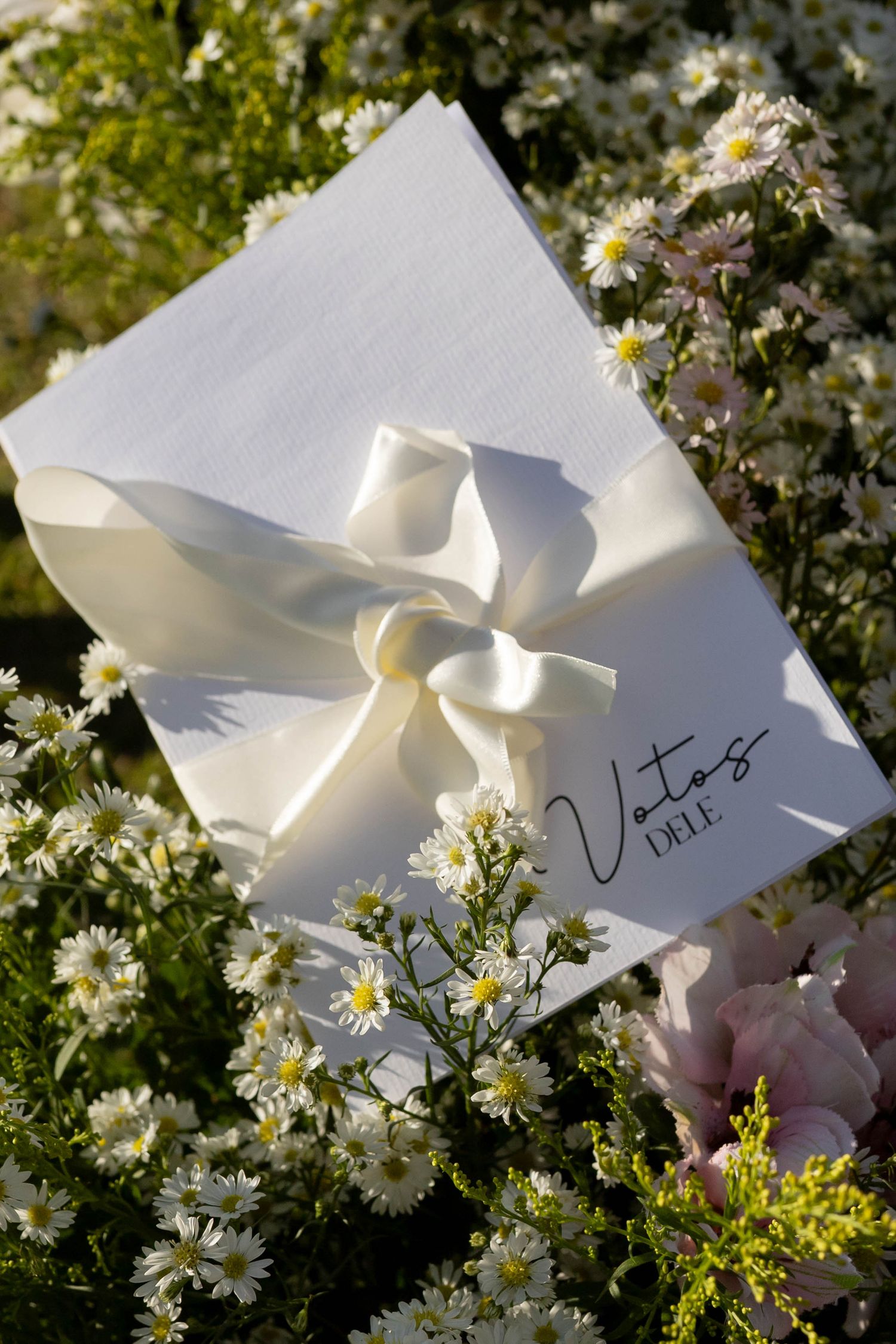 White gift box with satin ribbon bow labeled Votos DELE surrounded by delicate white daisy flowers.