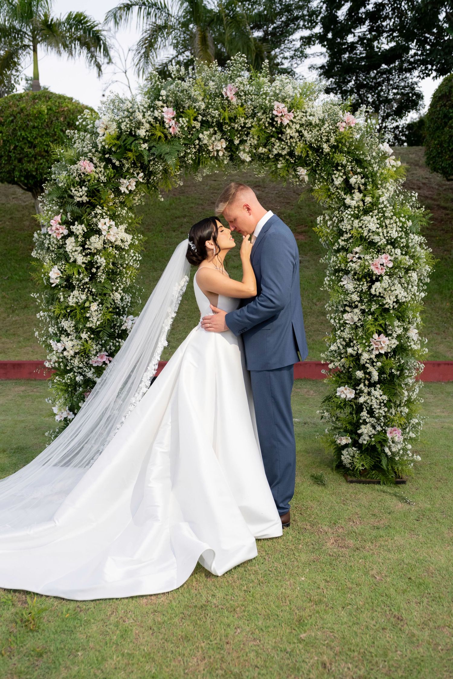 Bride and groom share a romantic kiss under a floral arch adorned with pink and white flowers at an outdoor wedding.