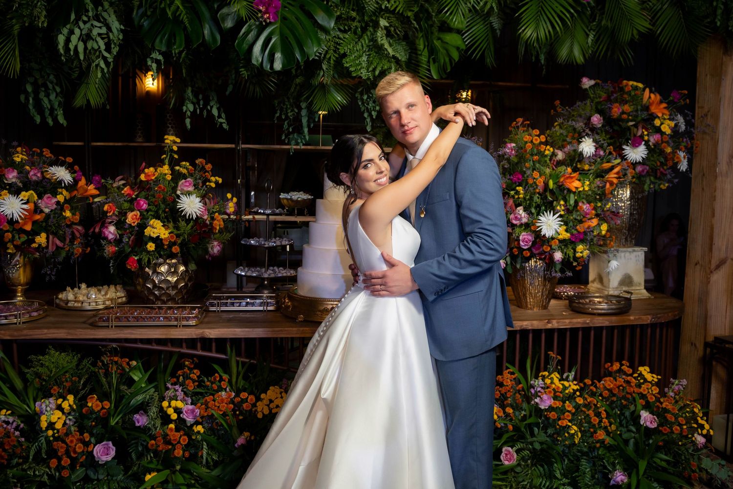 Bride and groom embrace in front of lush floral backdrop with colorful blooms at elegant wedding reception.