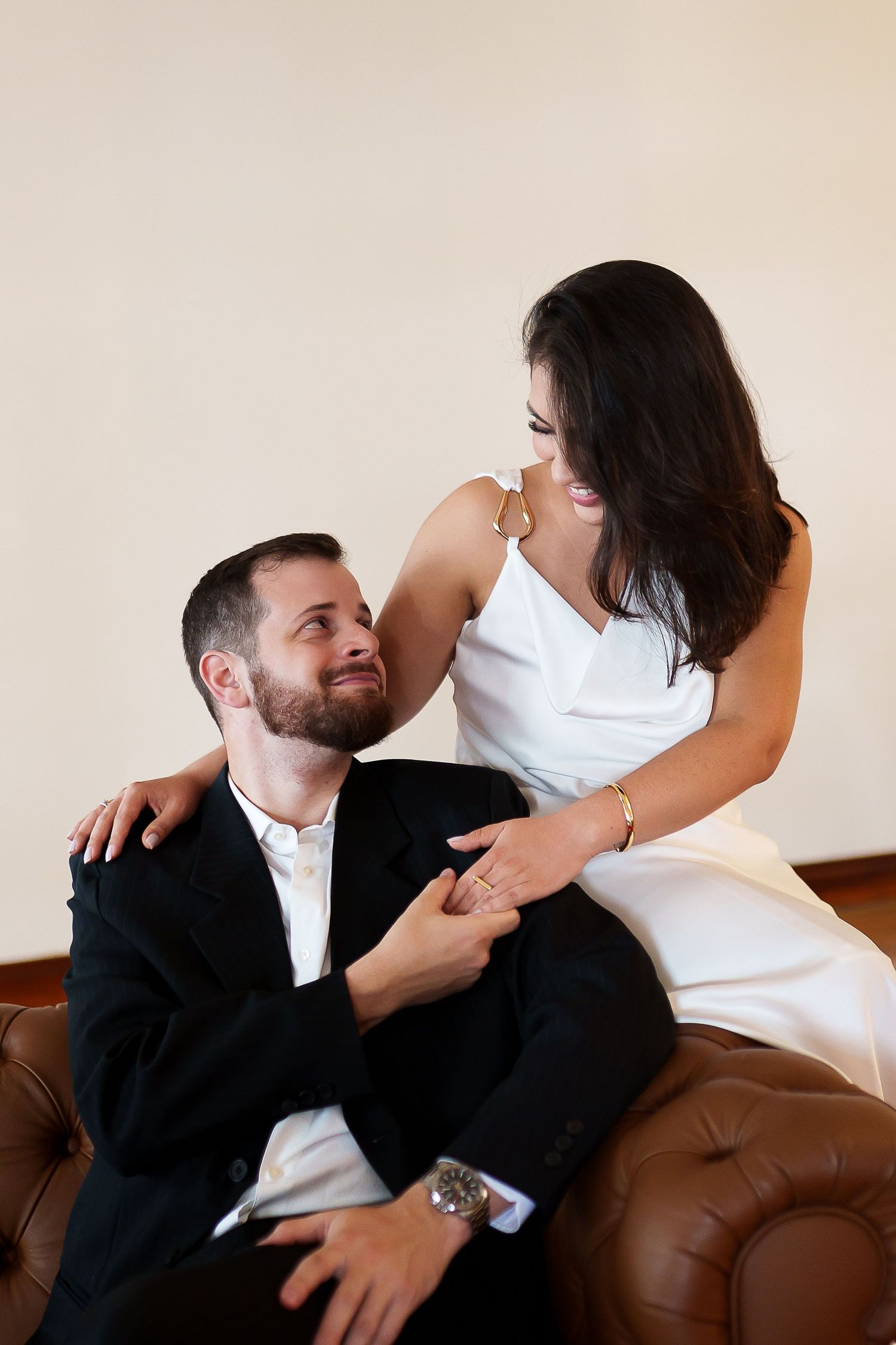 Elegant couple in formal wear sharing a romantic moment on a leather sofa, woman in white dress smiling at man in black suit.