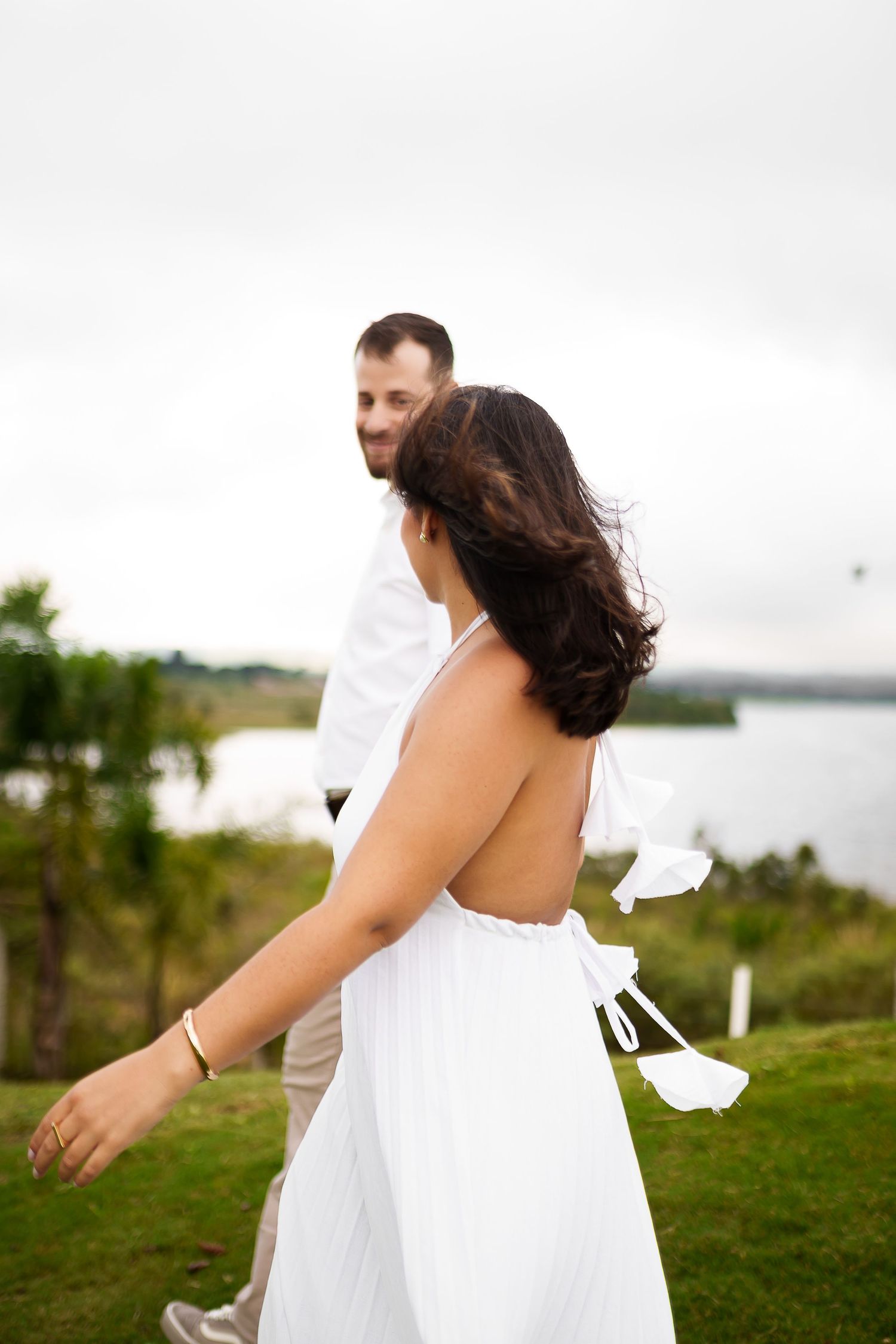 Couple in white outfits walking outdoors near a lake, woman in backless halter dress, man smiling behind her.
