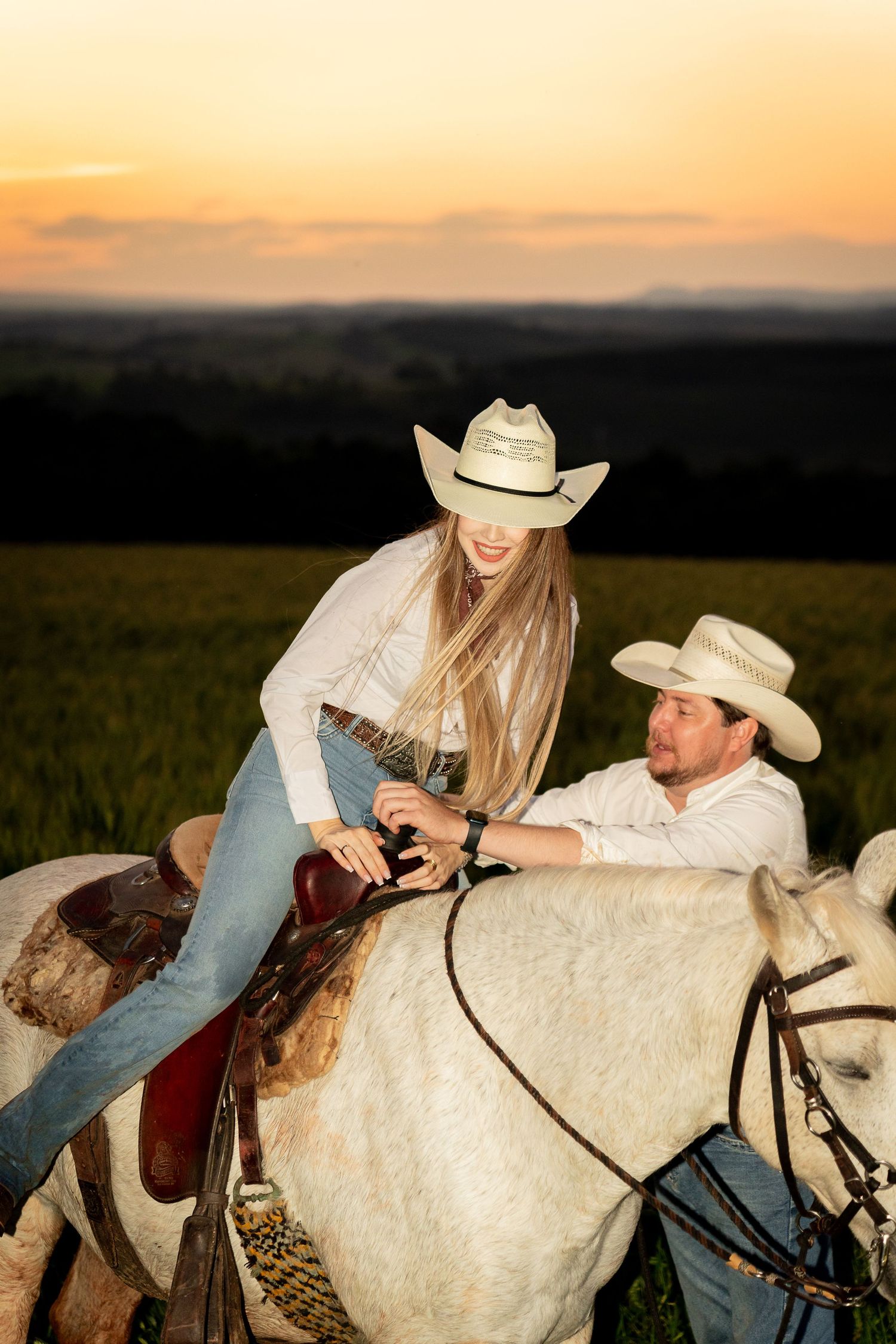 A smiling couple in cowboy hats on a white horse at sunset, with green fields in the background.