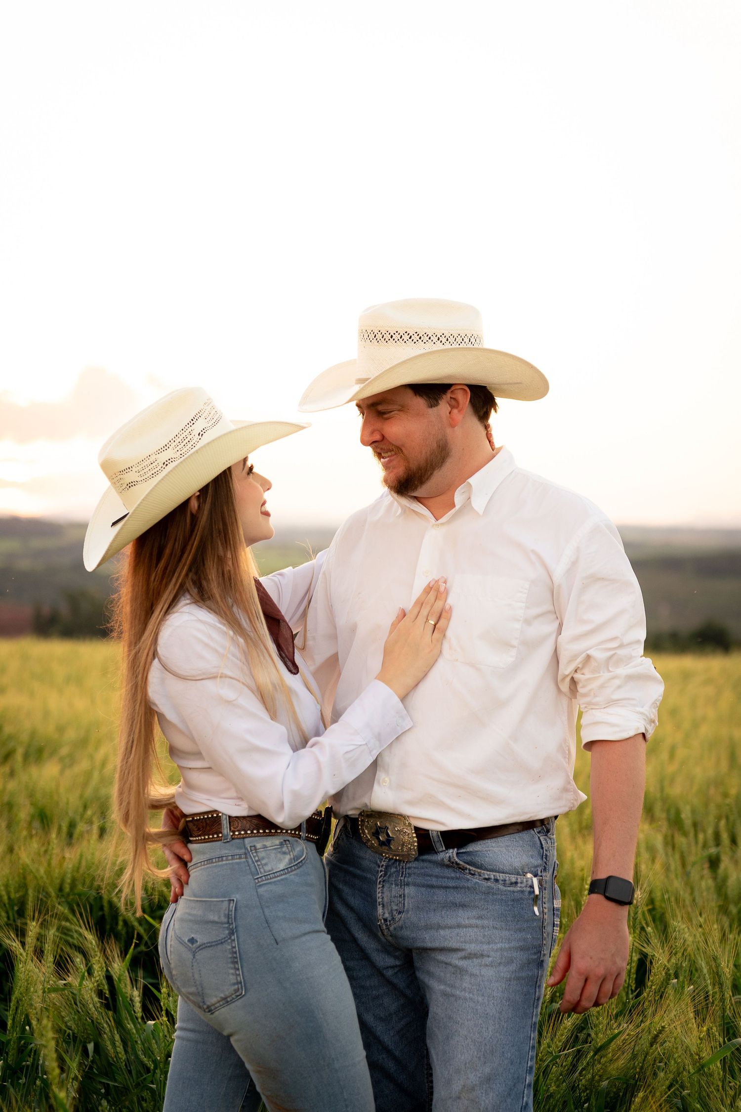 A romantic couple in matching white shirts, cowboy hats, and jeans embrace in a green field at golden hour.