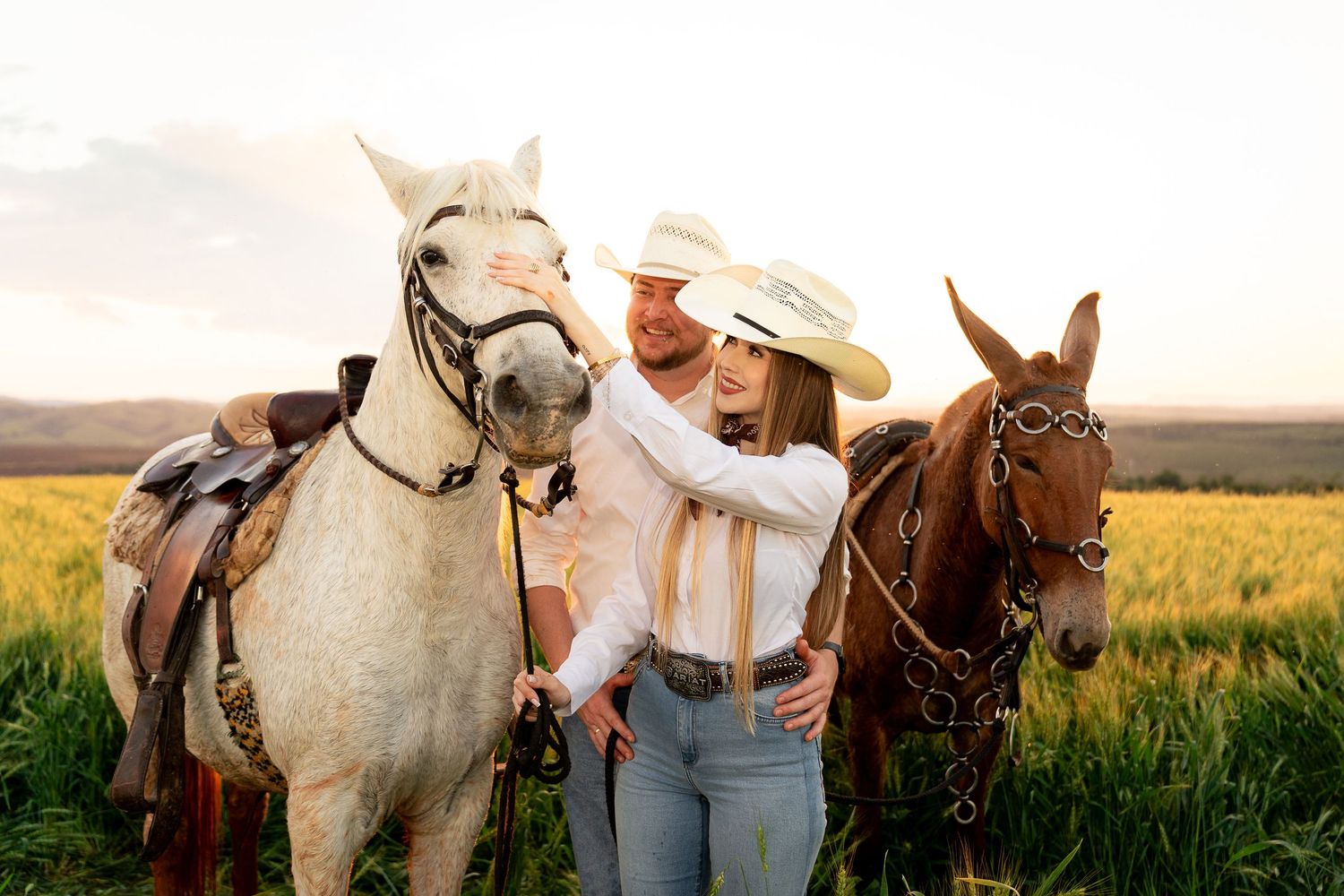 Couple in cowboy hats posing with a white and brown horse in a green field at sunset.