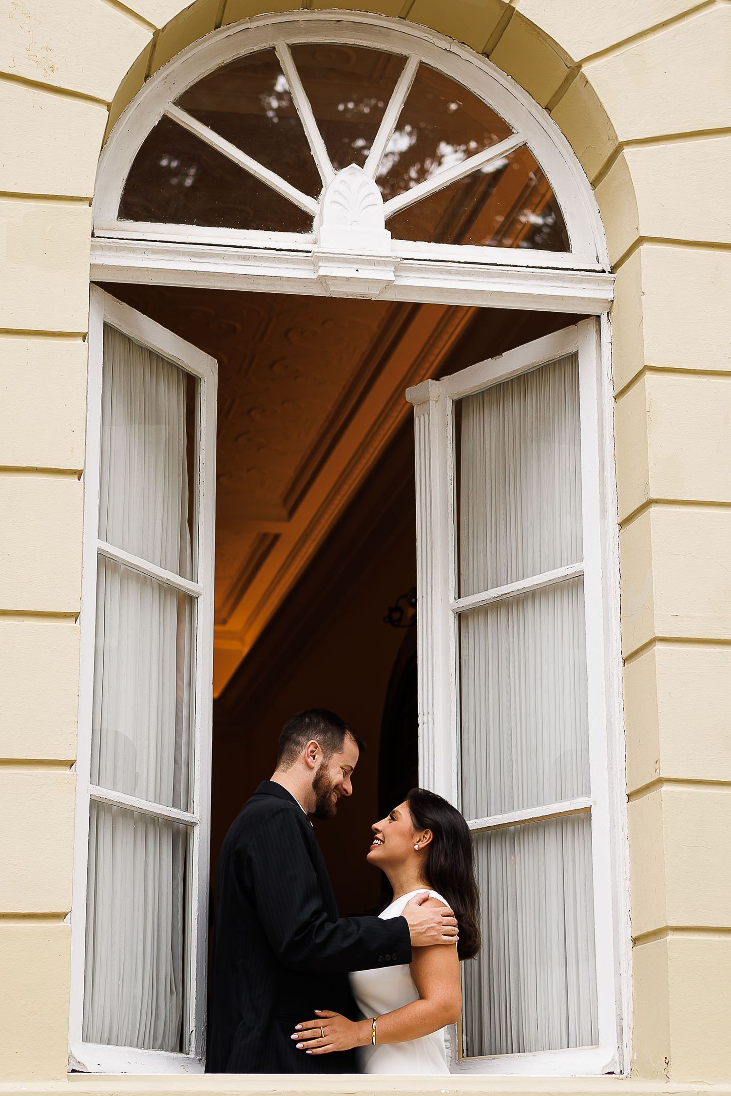 Bride and groom share a romantic moment in an arched window of an elegant yellow building.