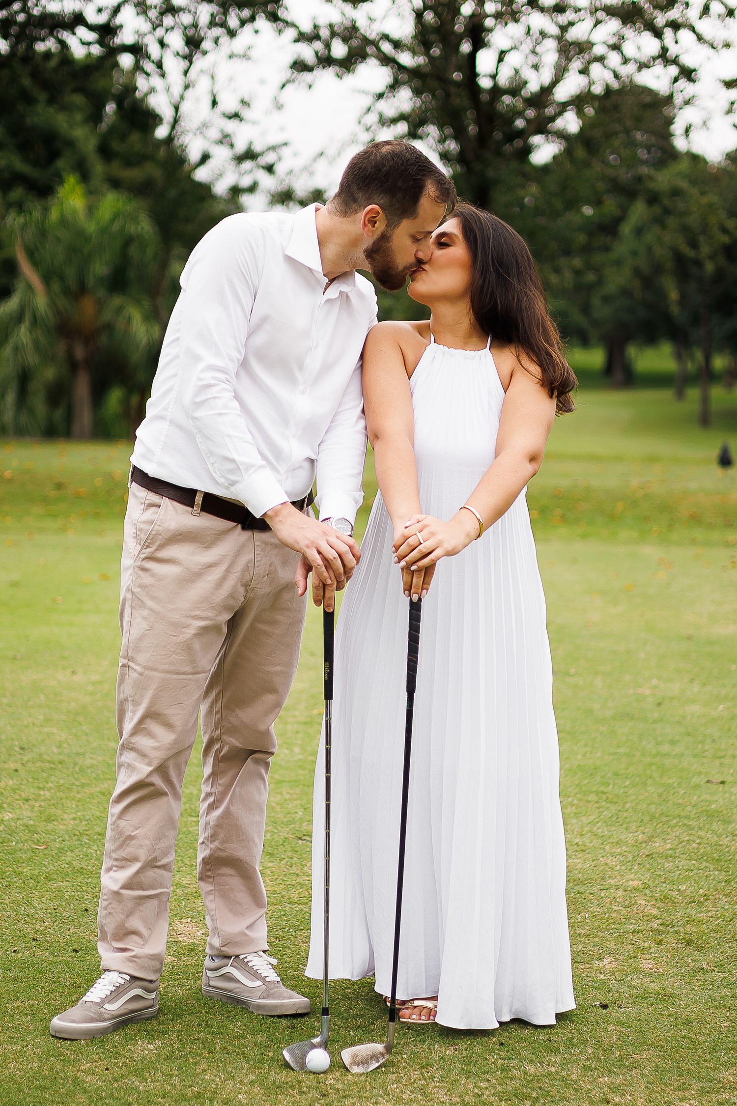 Couple sharing a romantic kiss on a golf course, woman in white dress and man in white shirt holding golf clubs.