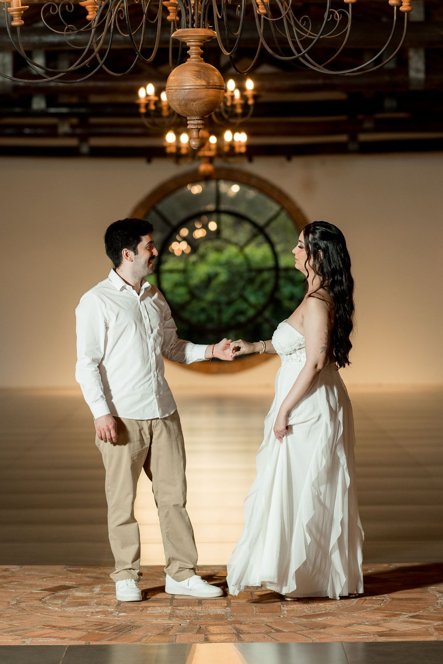 Couple holding hands in elegant venue with chandelier and round window backdrop, bride in white gown, groom in casual attire.